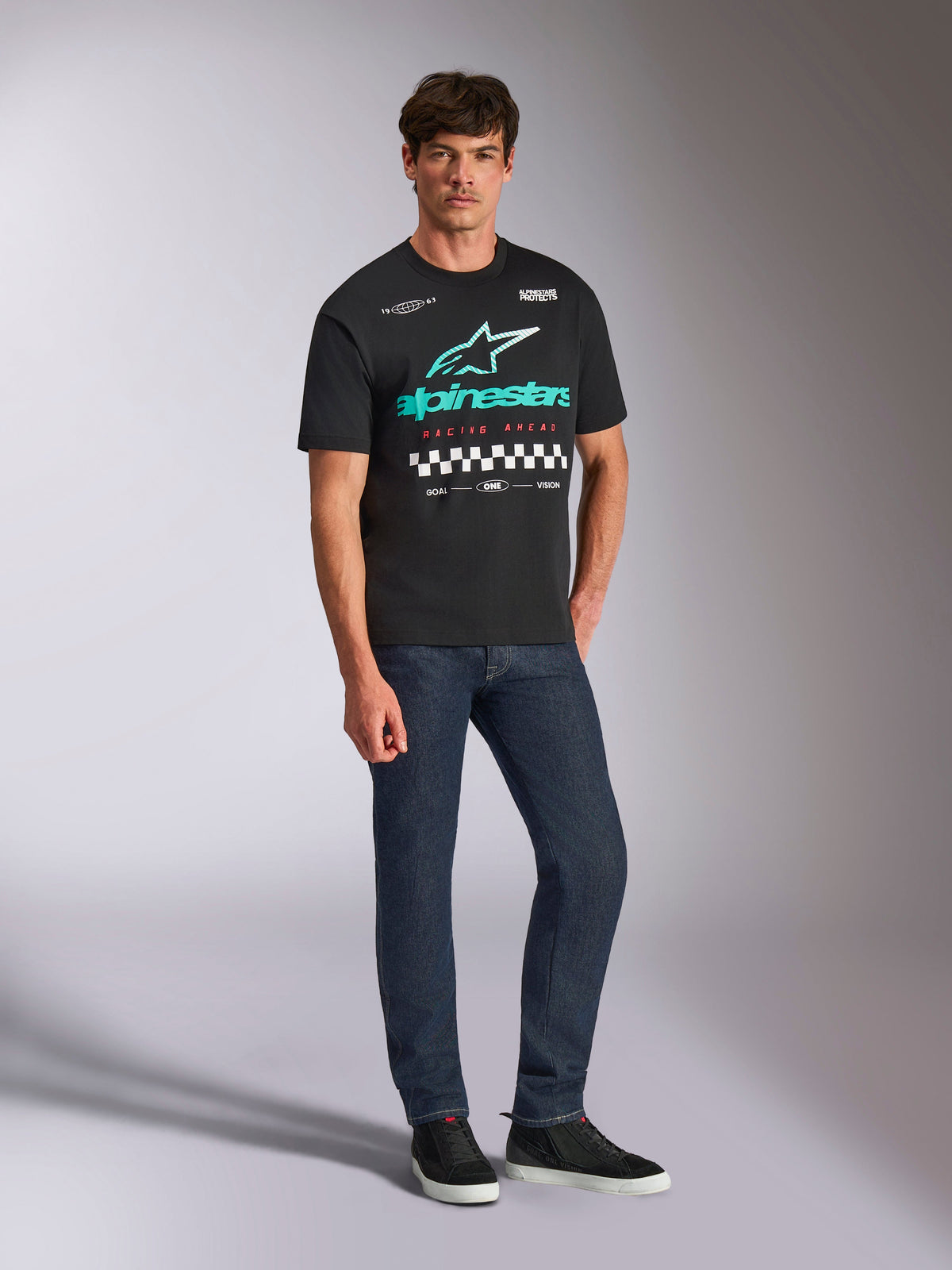 Alpinestars Aplomb Oversized CSF Tee, Short Sleeve T-Shirt, Black, Large teal and white Alpinestars logo with color gradient, Racing Ahead text and checkered pattern, worn with dark blue jeans and black sneakers, casual motorsport streetwear