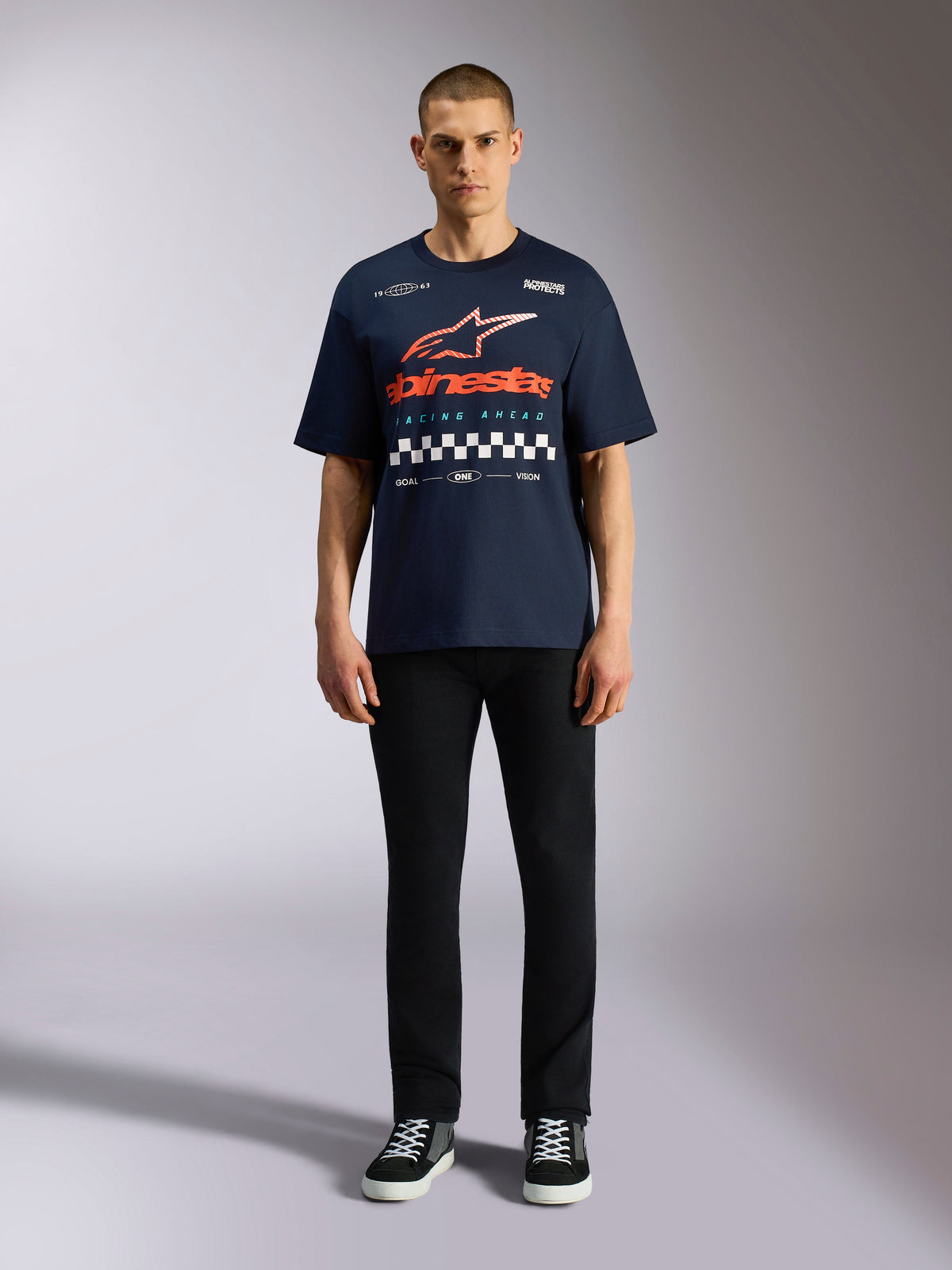 Alpinestars Aplomb Oversized CSF Tee, granatowy t-shirt z krótkim rękawem, duża grafika na klatce piersiowej z pomarańczowo-czerwonym gradientowym logo i wzorem szachownicy, model w czarnych spodniach i sneakersach, Casual Motorsport Streetwear