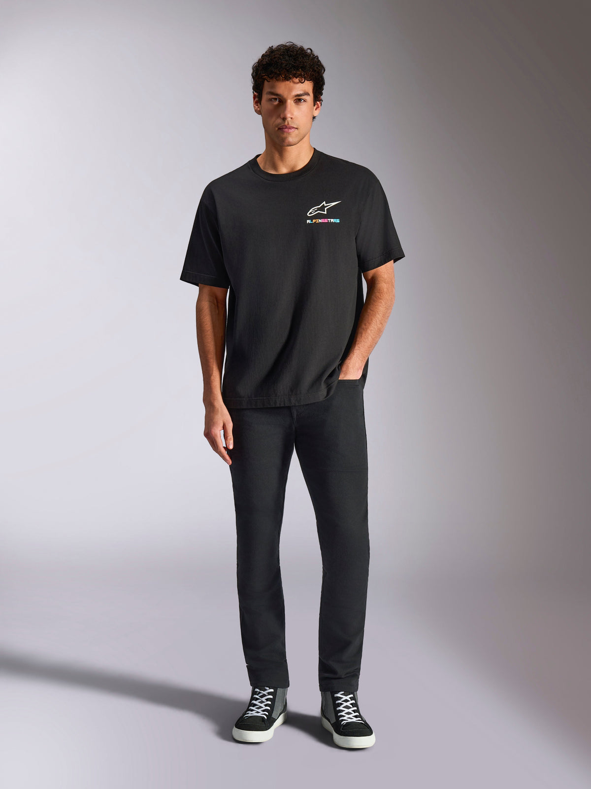 Alpinestars Wild Oversized CSF Tee, czarna koszulka z krótkim rękawem, noszona przez modela z czarnymi spodniami i sneakersami, białe logo Astars i żywy, wielokolorowy branding, casualowy streetwear motorsportowy