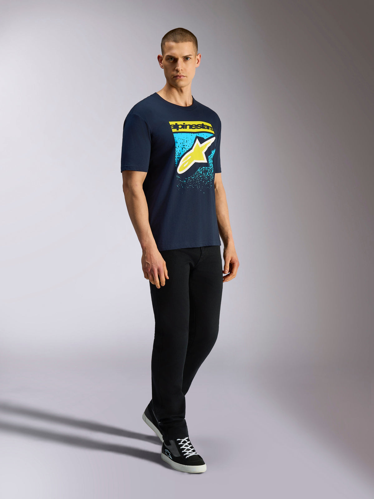 Alpinestars Noka CSF Tee, koszulka z krótkim rękawem, granatowa, żółte logo Astars z grafiką z niebieską gradacją MX, męski model w granatowej koszulce, czarnych spodniach i sneakersach, codzienna odzież uliczna w stylu motorsport