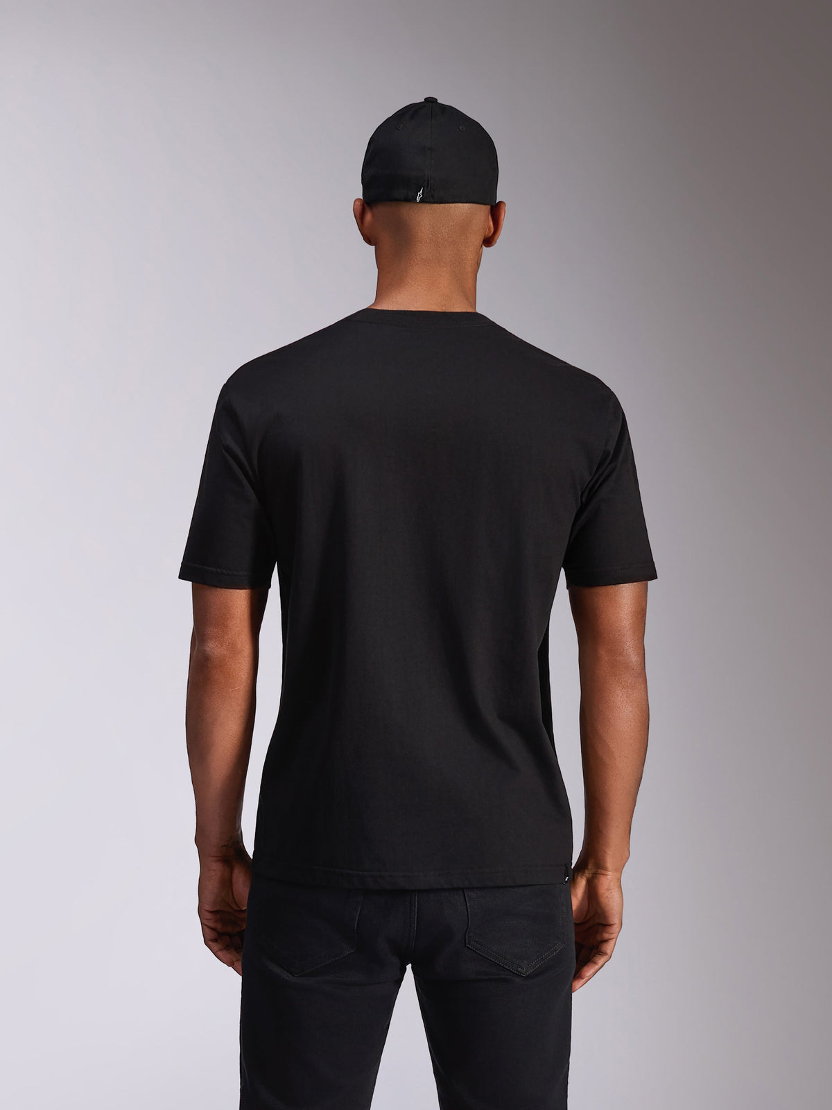 Alpinestars Ellipsoid CSF Tee, czarna koszulka z krótkim rękawem, widok z tyłu na modelu, zestawiona z czarną czapką i ciemnymi spodniami, codzienna odzież streetwearowa inspirowana sportami motorowymi