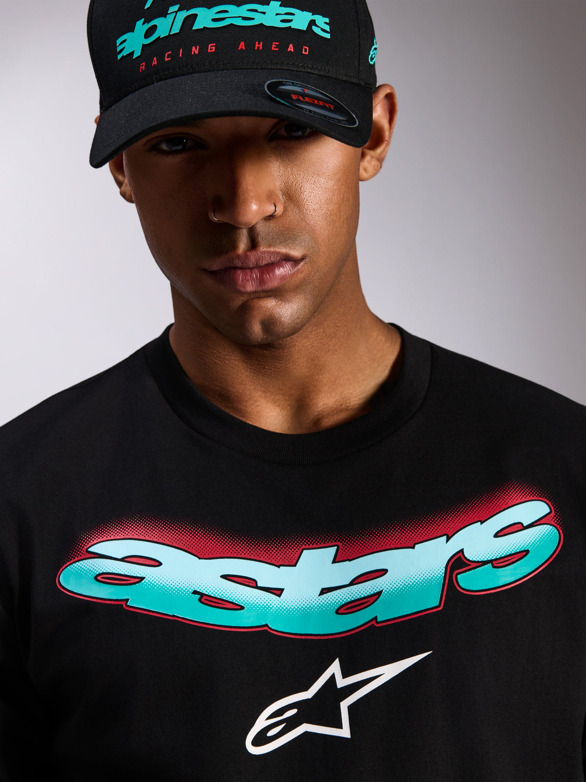 Alpinestars Ellipsoid CSF Tee, czarny t-shirt z krótkim rękawem, napis Astars z gradientem w kolorze morskim i białym oraz czerwonymi detalami typu halftone, białe logo Astars, noszony z dopasowaną czarną czapką Alpinestars, casualowa odzież streetwearowa w stylu motorsportowym
