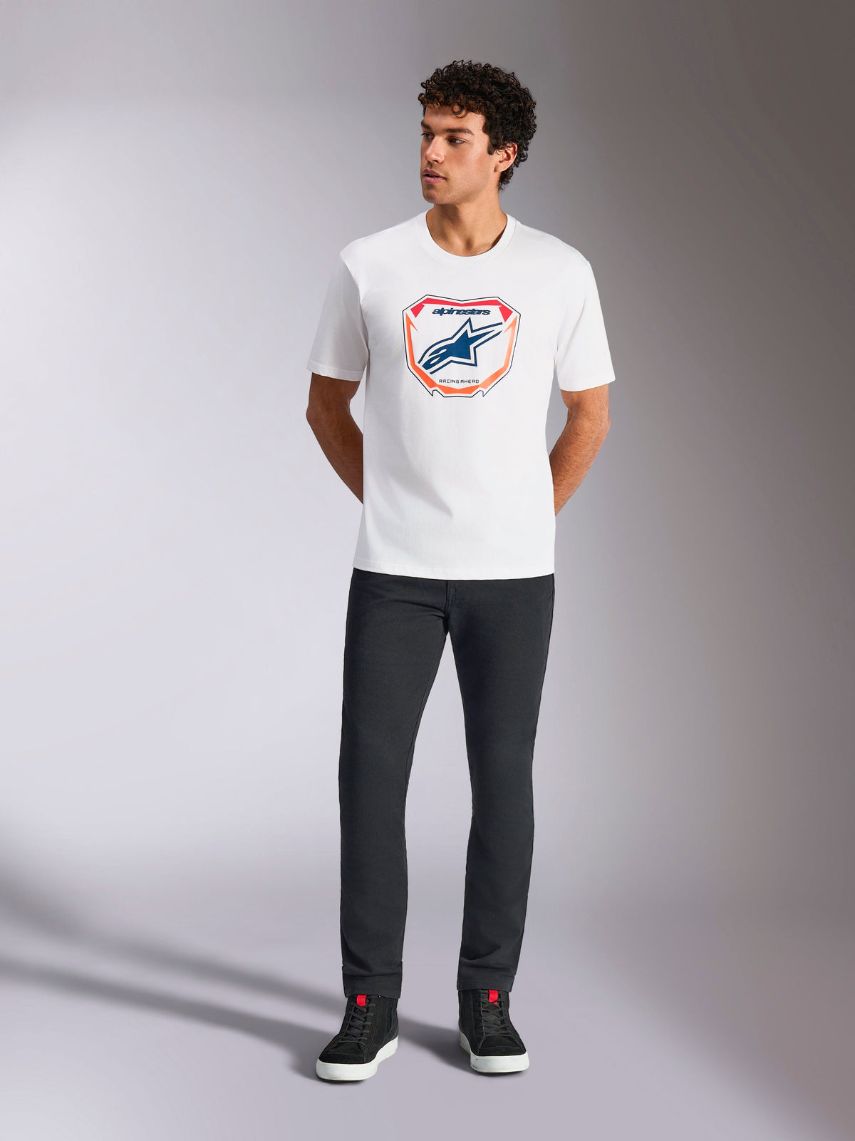 Alpinestars Plate CSF Tee, biała koszulka z krótkim rękawem, z grafiką płyty Astars MX na przodzie z pomarańczowymi i niebieskimi akcentami, prezentowana przez modela w czarnych spodniach i sneakersach, codzienna odzież w stylu motorsport