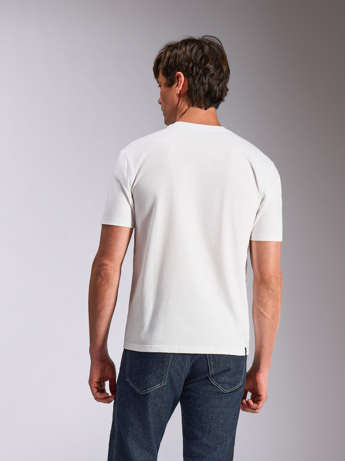 Alpinestars Abus CSF Tee, koszulka z krótkim rękawem, biała, widok z tyłu na modela w gładkiej białej koszulce i ciemnoniebieskich dżinsach, codzienna odzież lifestylowa inspirowana sportami motorowymi