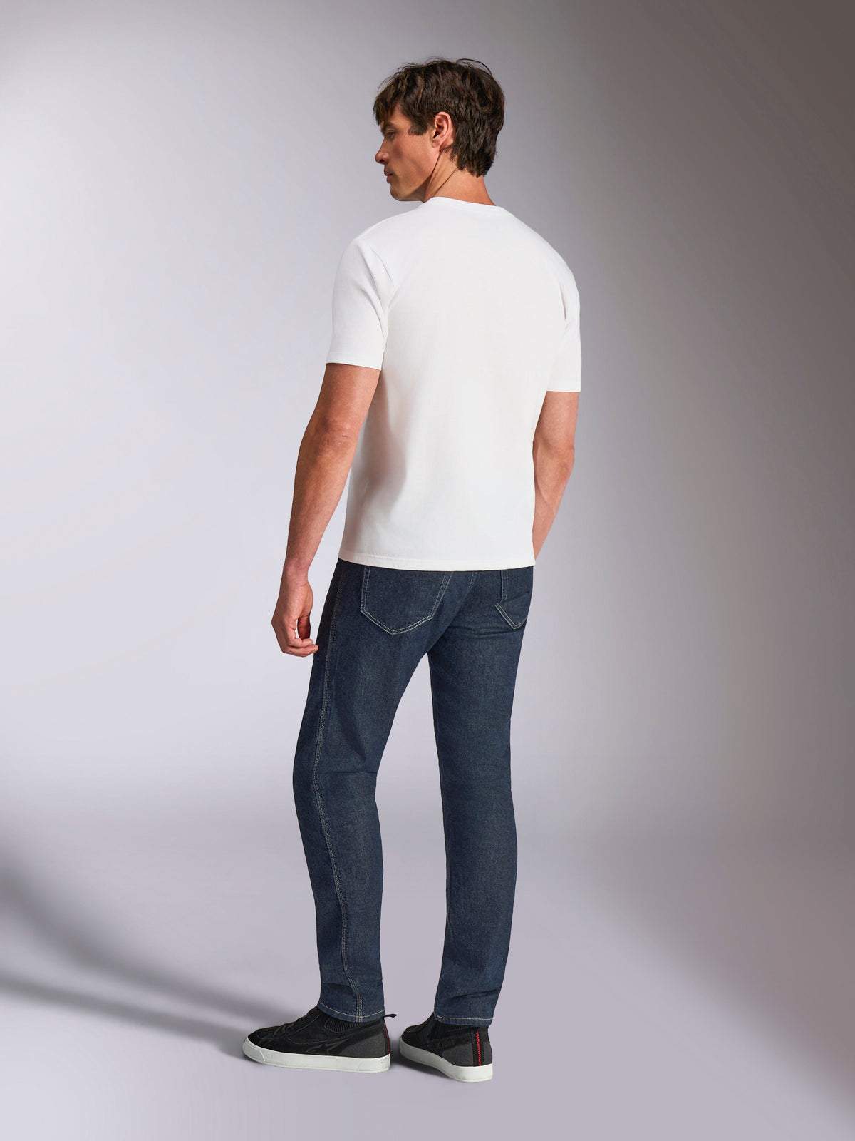 Alpinestars Abus CSF Tee, koszulka z krótkim rękawem, biała, widok z tyłu na modelu w ciemnoniebieskich dżinsach i czarnych sneakersach, codzienny design inspirowany sportami motorowymi