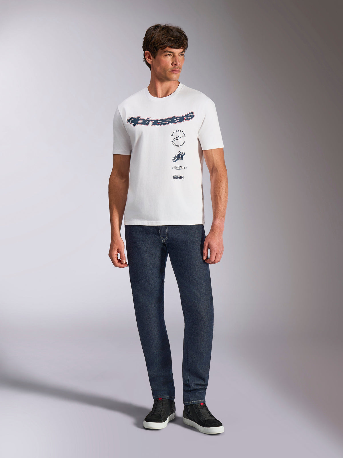 Alpinestars Abus CSF Tee, biała koszulka z krótkim rękawem, zakrzywione logo Alpinestars i wielowarstwowe grafiki na piersi, model w ciemnoniebieskich jeansach i czarnych sneakersach, casualowa odzież uliczna w stylu motorsport