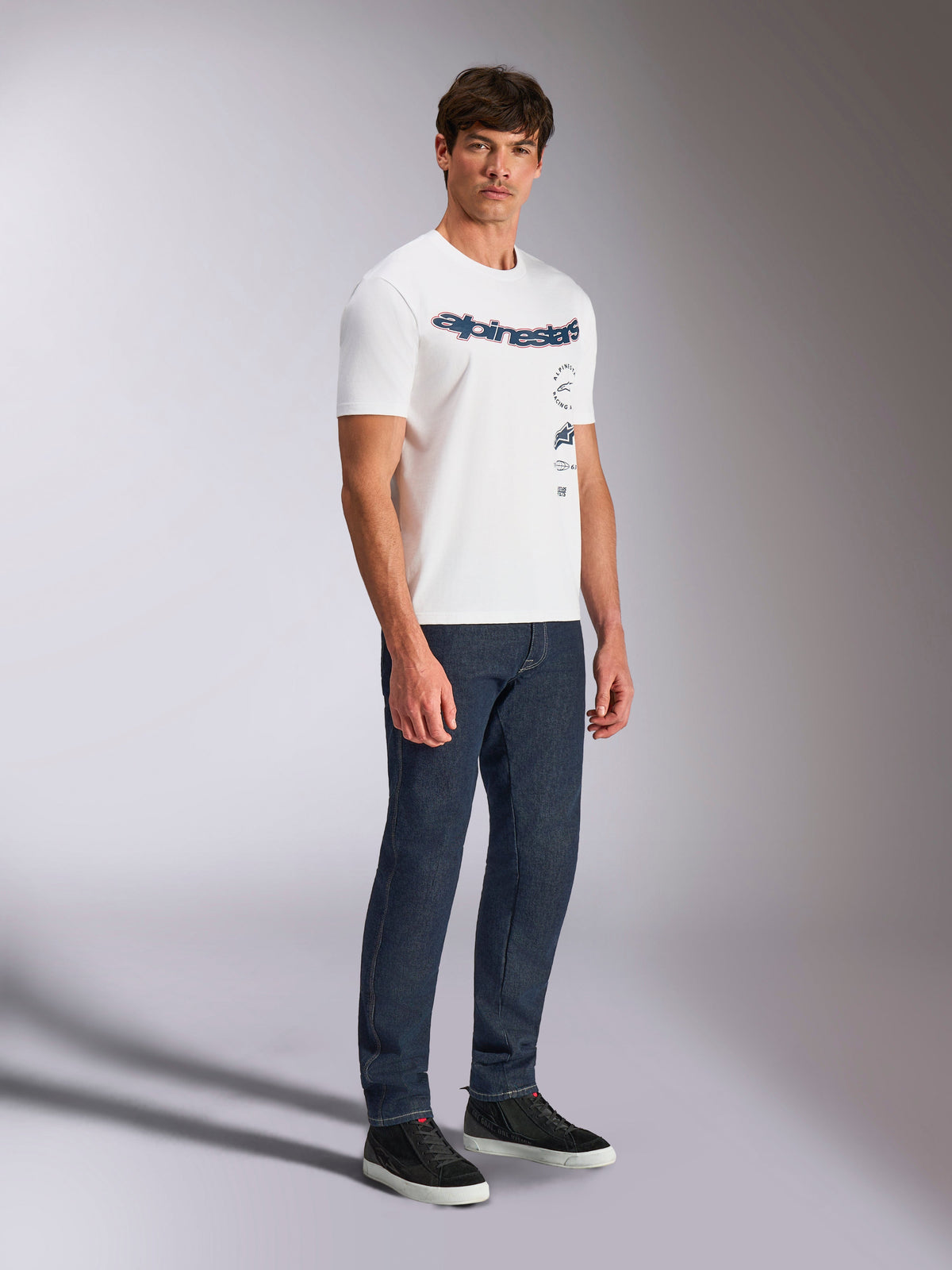 Alpinestars Abus CSF Tee, biała koszulka z krótkim rękawem, wielowarstwowe zakrzywione logo Alpinestars, noszona przez modela z ciemnoniebieskimi dżinsami i czarnymi sneakersami, codzienna odzież uliczna w stylu motorsportowym