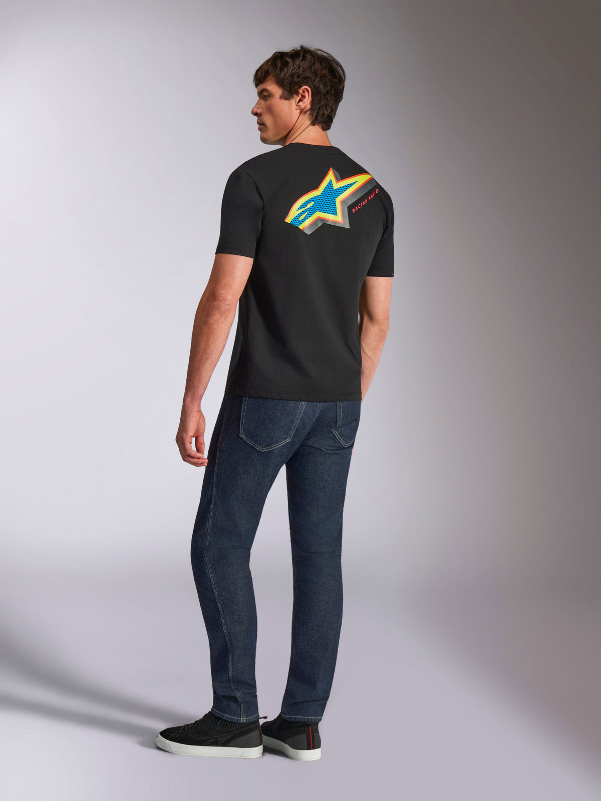 Alpinestars Angler CSF Tee, koszulka z krótkim rękawem, czarna, z dużym wyrazistym wielokolorowym logo Astars na plecach z niebieskimi, żółtymi i czerwonymi perforowanymi detalami, noszona z ciemnoniebieskimi dżinsami, casualowa odzież uliczna w stylu motorsport