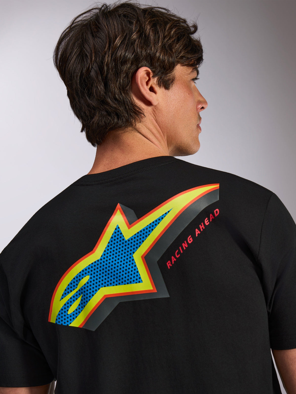 Alpinestars Angler CSF Tee, czarna koszulka z krótkim rękawem, duże wielokolorowe logo Astars na plecach z niebieskim perforowanym wypełnieniem, żółtymi i czerwonymi obramowaniami, napis „RACING AHEAD”, codzienna odzież motorsportowa