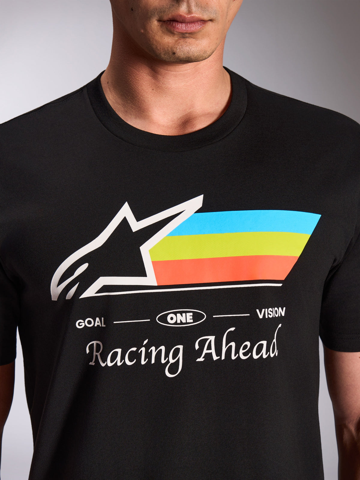 Alpinestars Trifecta CSF Tee, czarna koszulka z krótkim rękawem, zbliżenie na grafikę z przodu z białym logo Astars i trójkolorowymi niebieskimi, zielonymi i pomarańczowymi paskami, napis „Racing Ahead”, casualowa odzież motorsportowa