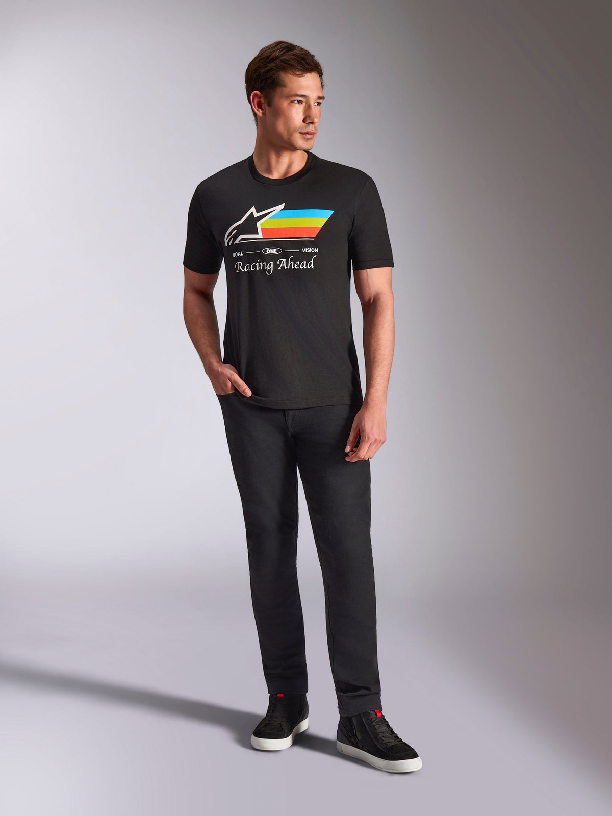 Alpinestars Trifecta CSF Tee, koszulka z krótkim rękawem, czarna, z białym logo Astars, błękitnymi, żółtymi i pomarańczowymi pasami oraz napisem „Racing Ahead”, prezentowana przez modela w czarnych spodniach i sneakersach, styl casual motorsport streetwear