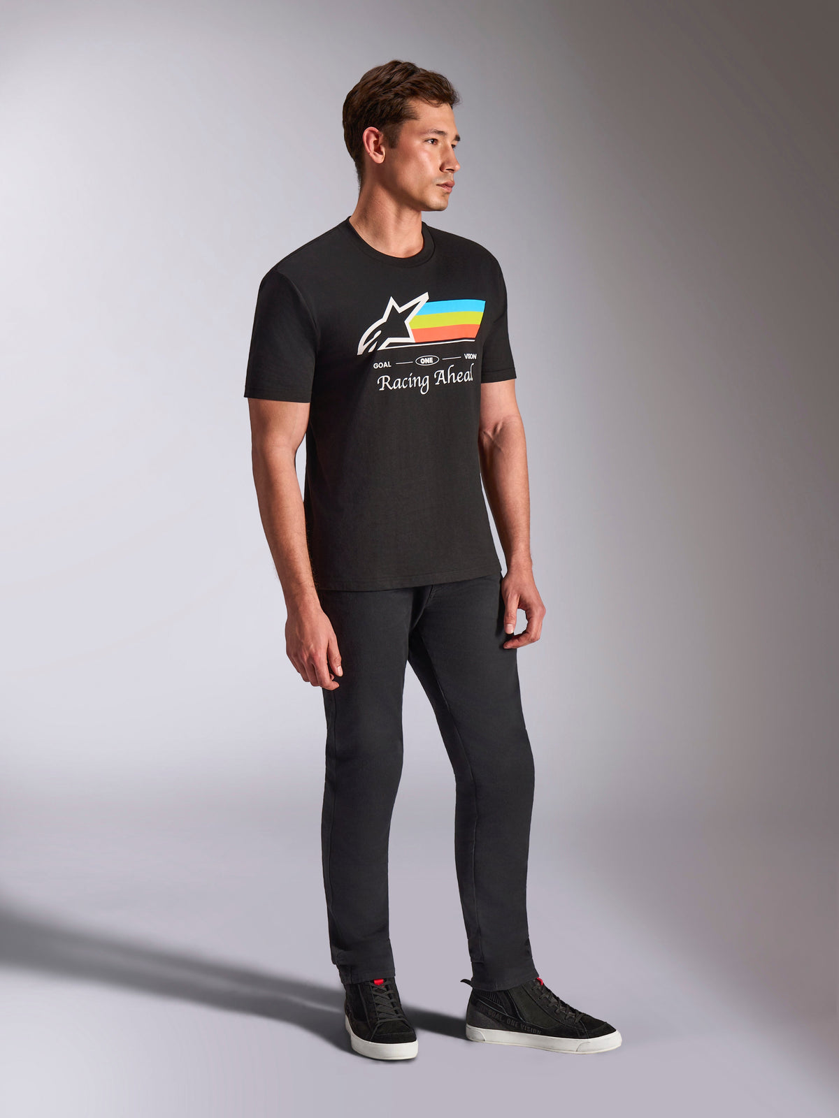 Alpinestars Trifecta CSF Tee, koszulka z krótkim rękawem, czarna, białe logo Astars z niebieskimi, żółtymi i pomarańczowymi paskami oraz grafiką „Racing Ahead”, prezentowana przez modela w czarnych spodniach i sneakersach, codzienna odzież lifestylowa inspirowana sportami motorowymi