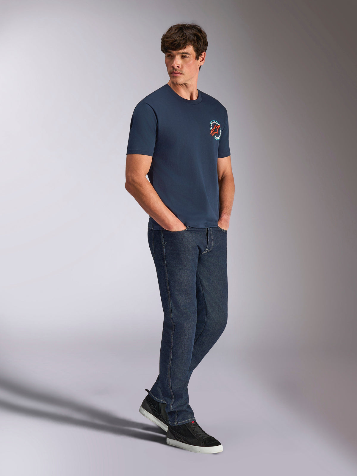 Alpinestars Aorbit CSF Tee, Short Sleeve T-Shirt, Navy, featuring circular orange and white Astars logo with Always Fast Made to Last graphic, worn by a man with dark denim jeans and black sneakers, casual motorsport-inspired streetwear