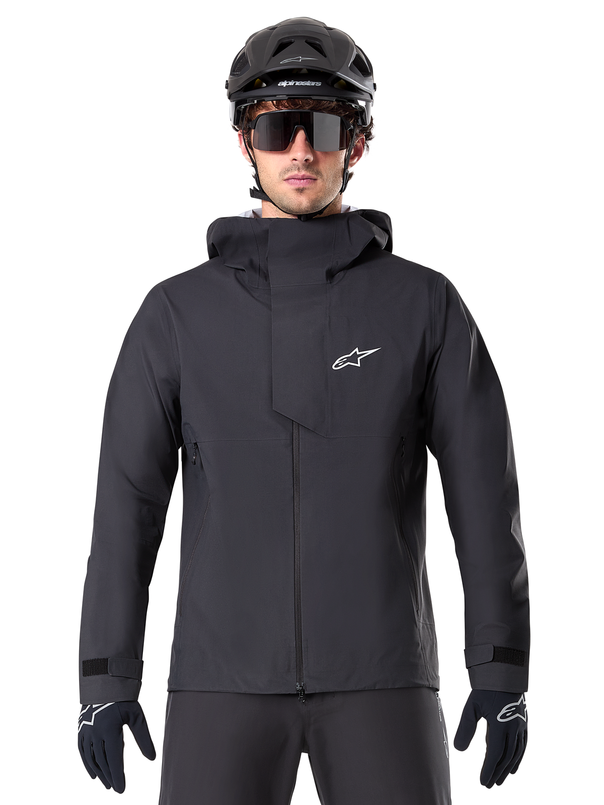 Alpinestars A-Dura Elite Waterproof Jacket, czarna kurtka MTB, wysoki kołnierz z kapturem, przedni zamek z ochronną patką na klatce piersiowej i białym logo, noszona z czarnym kaskiem i rękawiczkami rowerowymi, techniczna ochrona przed warunkami atmosferycznymi na szlaku