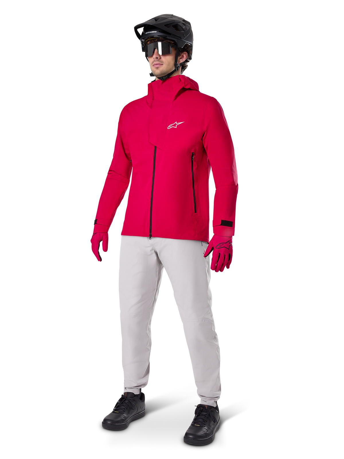 Rowerzysta ubrany w wodoodporną kurtkę Alpinestars A-Dura Elite w kolorze Red Fluo, jasnoszare spodnie techniczne, czerwone rękawiczki, czarny kask MTB z goglami i czarne buty; kompletny, wysokowydajny strój do kolarstwa górskiego.