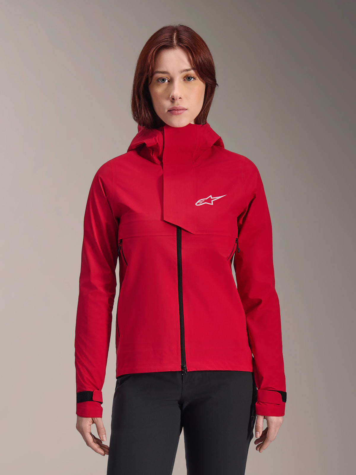 Alpinestars Stella A-Dura Elite Waterproof Jacket, damska kurtka rowerowa MTB, czerwona fluo, z kapturem i asymetryczną przednią klapą, białe logo Alpinestars na klatce piersiowej, regulowane czarne mankiety, prezentowana na modelce w czarnych spodniach