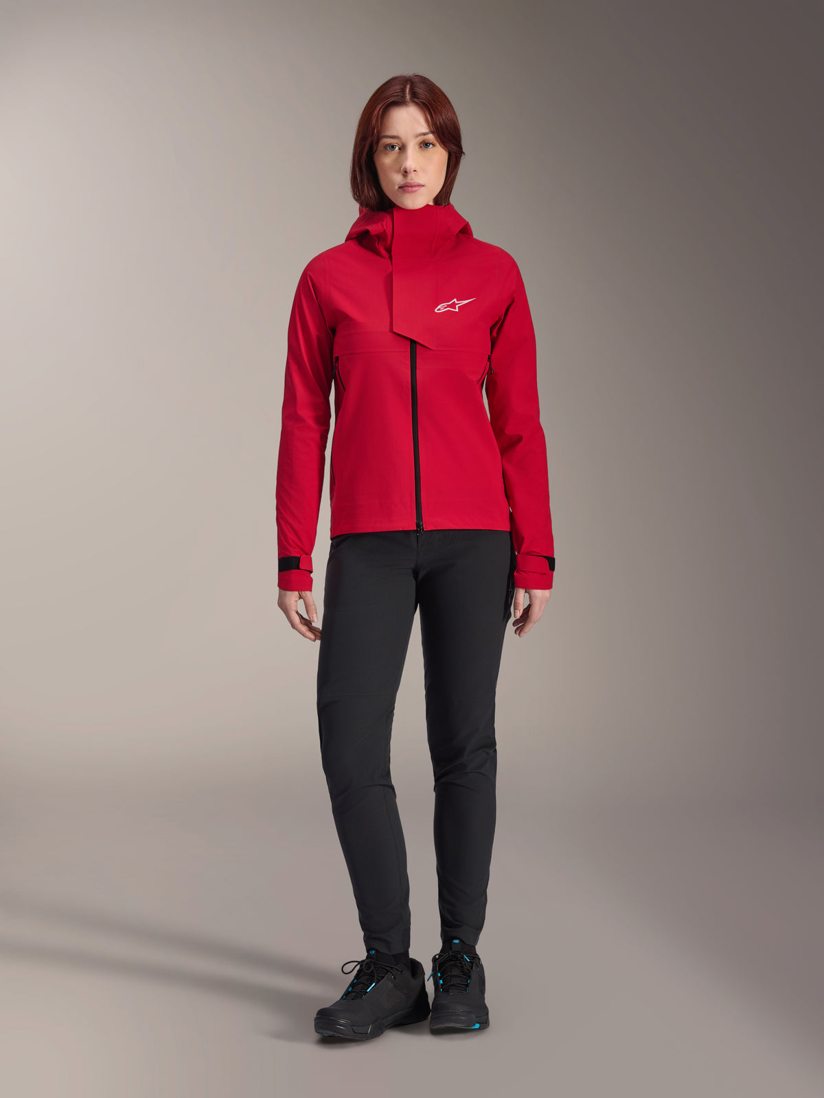 Wodoodporna kurtka Alpinestars Stella A-Dura Elite, damska kurtka MTB, kolor Red Fluo, noszona z czarnymi spodniami i butami, z kapturem z wysokim kołnierzem i białym logo Alpinestars, techniczna odzież na rower górski