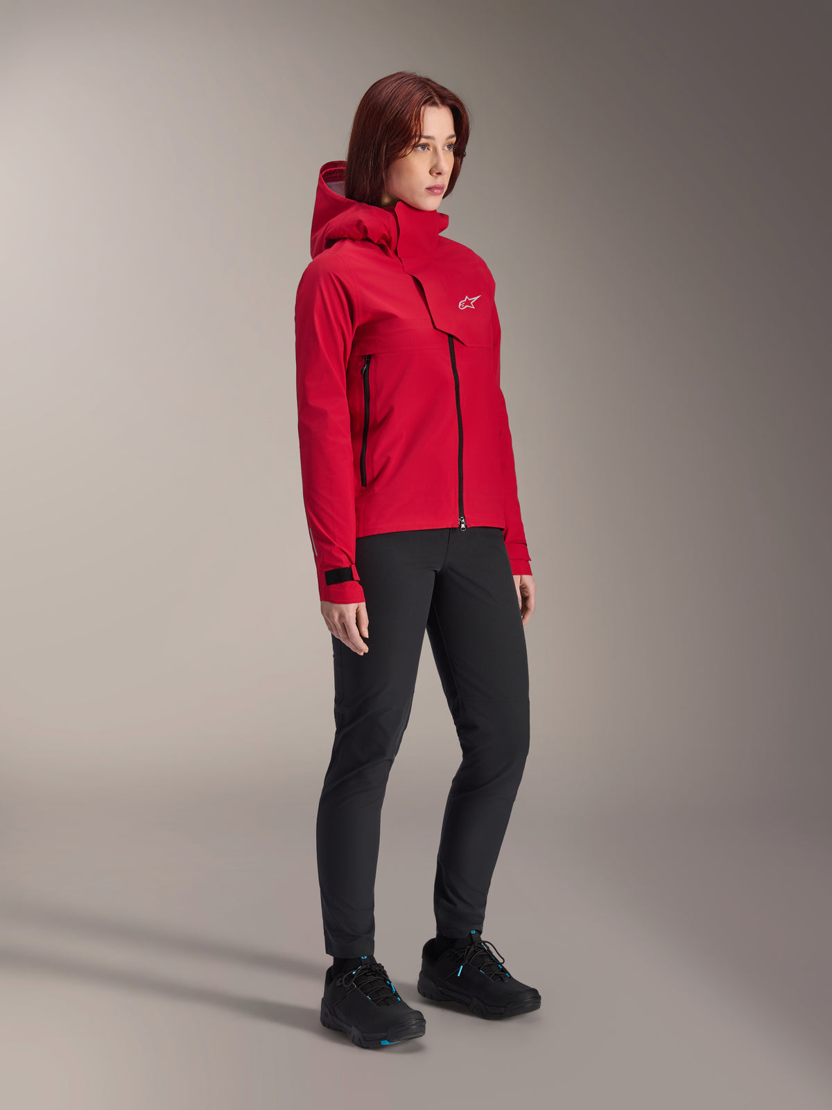 Damska kurtka Alpinestars Stella A-Dura Elite Waterproof, czerwony fluo, przeciwdeszczowa kurtka z kapturem na rower górski z białym logo Astars na klapie na klatce piersiowej, noszona z czarnymi spodniami i czarnymi butami rowerowymi MTB, techniczna odzież wyczynowa do jazdy po szlakach