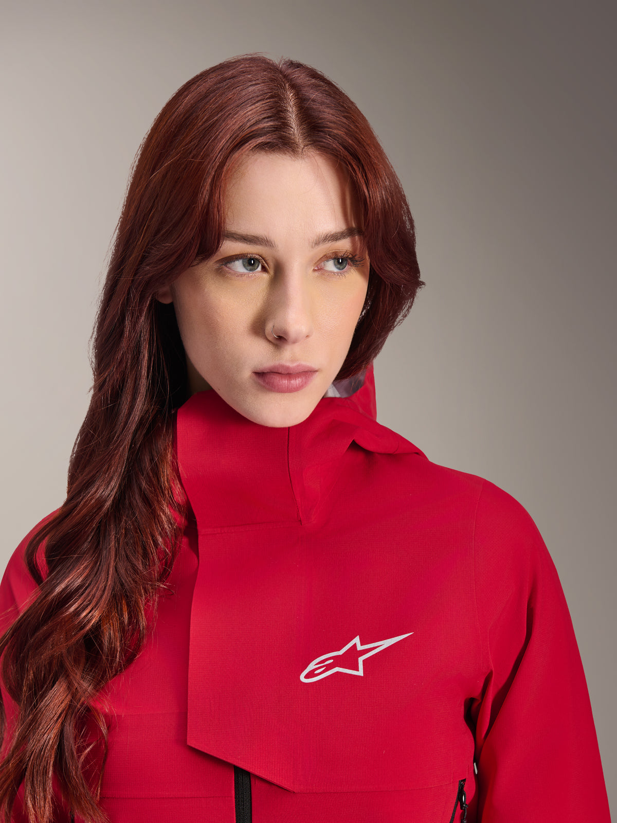 Alpinestars Stella A-Dura Elite Waterproof Jacket, Red Fluo, noszona przez kobietę z długimi rudymi włosami, techniczna 3-warstwowa wodoodporna kurtka z wysokim kołnierzem i białym logo, odzież outdoorowa i rowerowa MTB o wysokiej wydajności