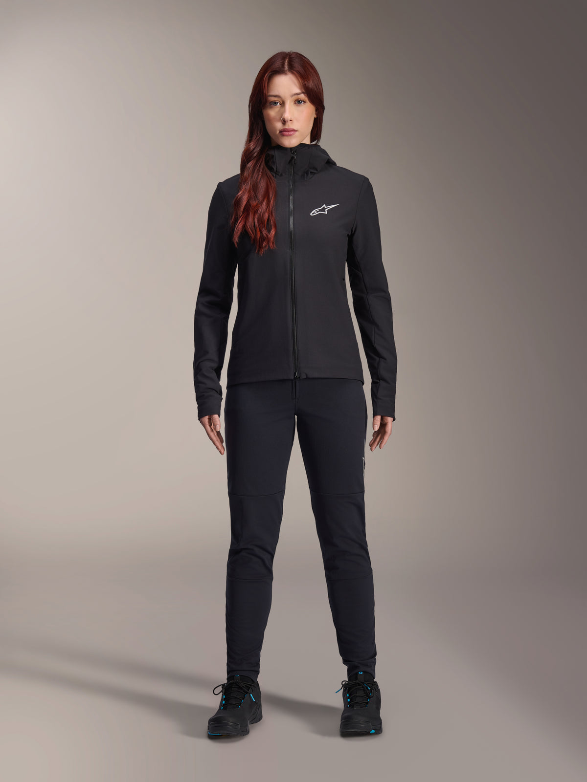 Kobieta w kurtce Alpinestars Stella A-Dura Thermal, czarna lekka ocieplana kurtka rowerowa z kapturem i białym logo Astars na piersi, w czarnych spodniach i butach sportowych