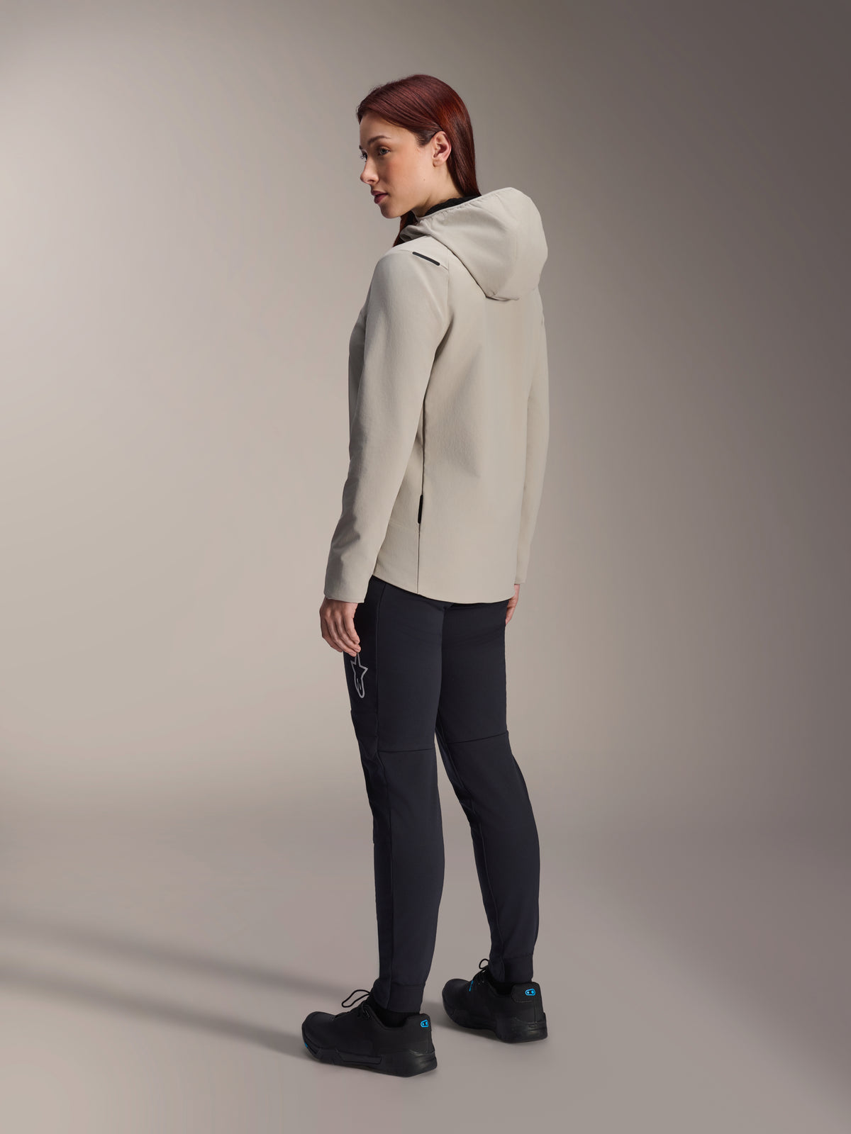 Alpinestars Stella A-Dura Thermal Jacket, szara ocieplana kurtka rowerowa z kapturem, widok z tyłu pod kątem trzech czwartych na kobietę w czarnych spodniach MTB, ujęcie studyjne, regularny krój i detale kieszeni