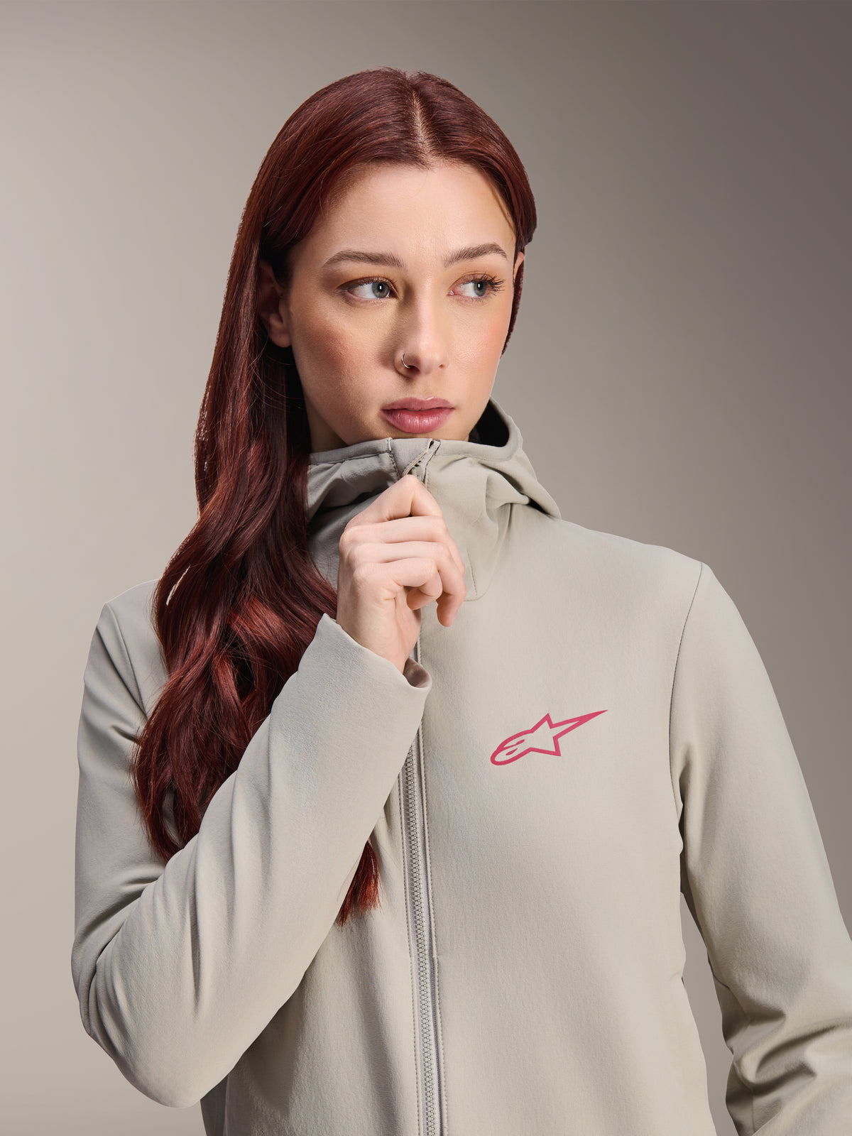 Damska kurtka termiczna Alpinestars Stella A-Dura, kolor szary Titanium z różowym logo gwiazdy Alpinestars, modelka z długimi rudymi włosami zapinająca kurtkę z kapturem, lekka ocieplana odzież rowerowa na zimną pogodę