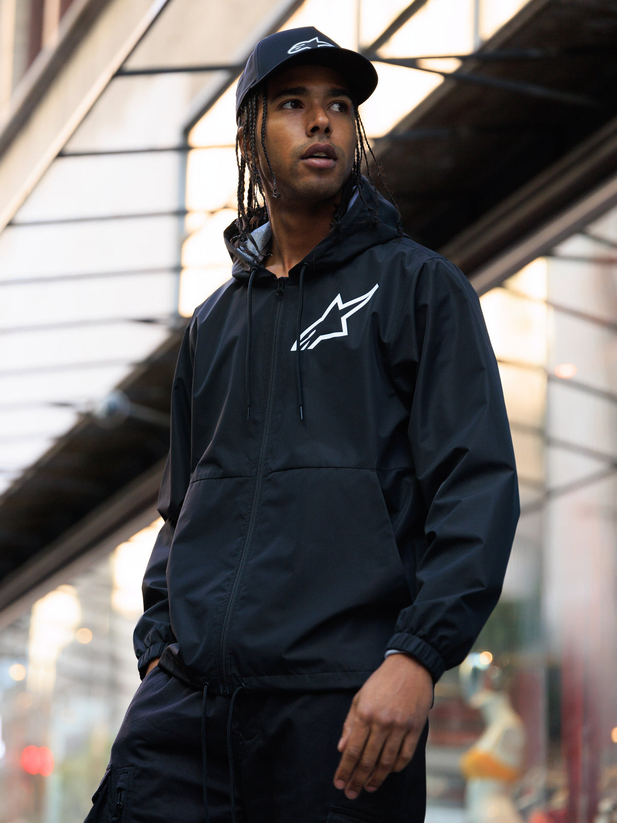 Kurtka Alpinestars Speeder Chromium Windbreak, czarna wiatrówka z kapturem z białym logo na piersi, noszona przez mężczyznę w pasującej czarnej czapce Alpinestars w miejskim otoczeniu, casualowy streetwear w stylu motorsport