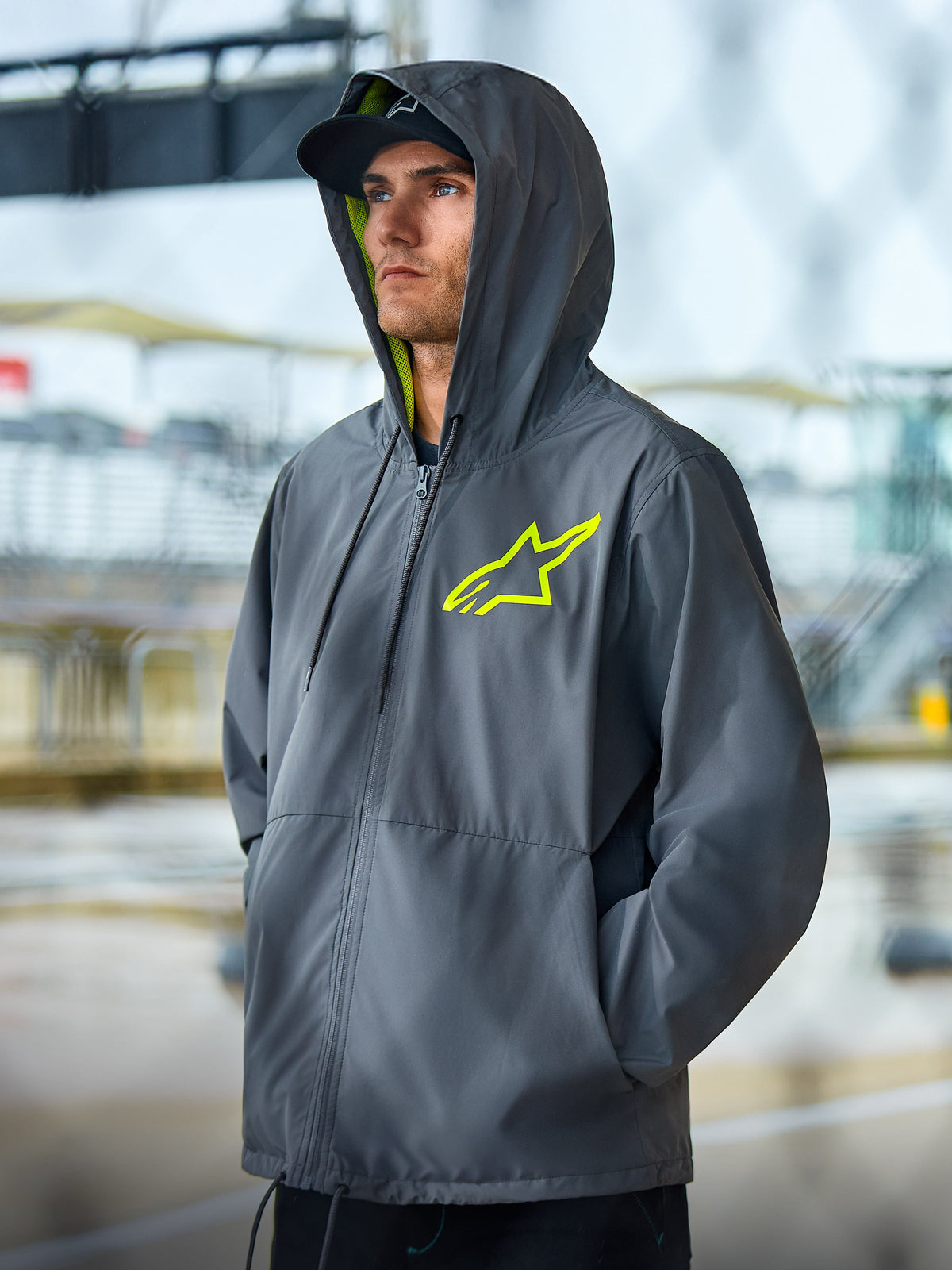 Alpinestars Speeder Chromium Windbreak Jacket, grafitowa kurtka typu lifestyle z jaskrawożółtymi akcentami, fason z kapturem, żółtą podszewką i logo na piersi, casualowy streetwear inspirowany motorsportem, noszona przez modela w plenerze