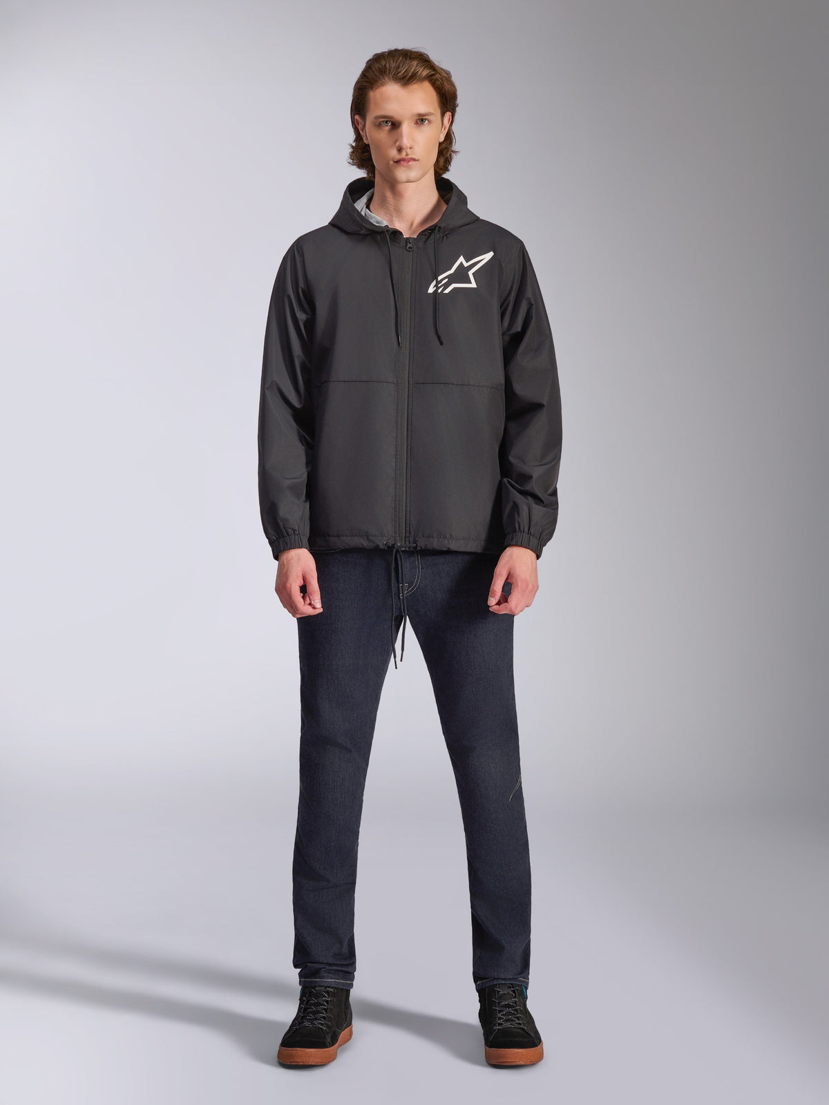 Kurtka wiatrówka Alpinestars Speeder Chromium, czarna z białym logo Astars, noszona przez modela z ciemnoniebieskimi jeansami i czarnymi sneakersami, z kapturem i zamkiem z przodu, casualowy streetwear inspirowany sportami motorowymi