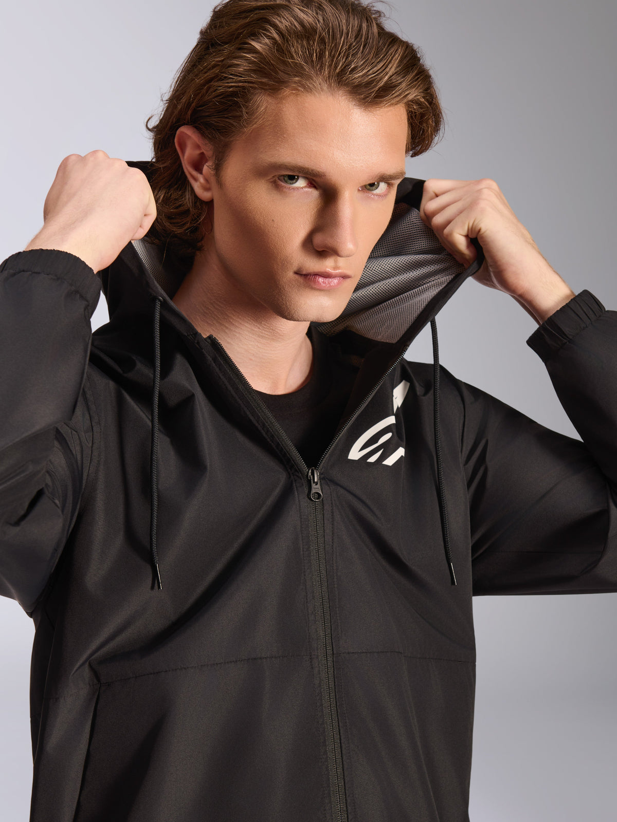 Kurtka przeciwwiatrowa Alpinestars Speeder Chromium, czarna, noszona przez modela trzymającego kaptur, posiada białe logo na piersi, siateczkową podszewkę kaptura i zamek na całej długości, codzienna odzież uliczna inspirowana motorsportem