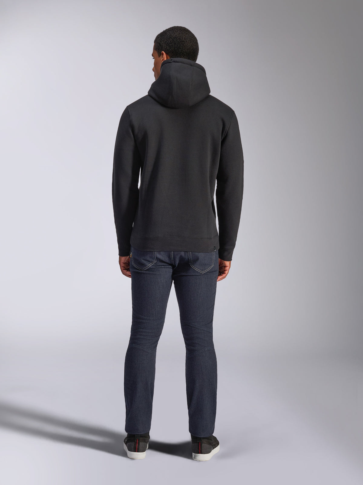 Alpinestars Blaze V3 Hoodie, codzienna odzież typu streetwear, czarna, widok z tyłu polarowej bluzy z kapturem, noszona z ciemnoniebieskimi dżinsami i czarnymi butami, czysty design inspirowany sportami motorowymi