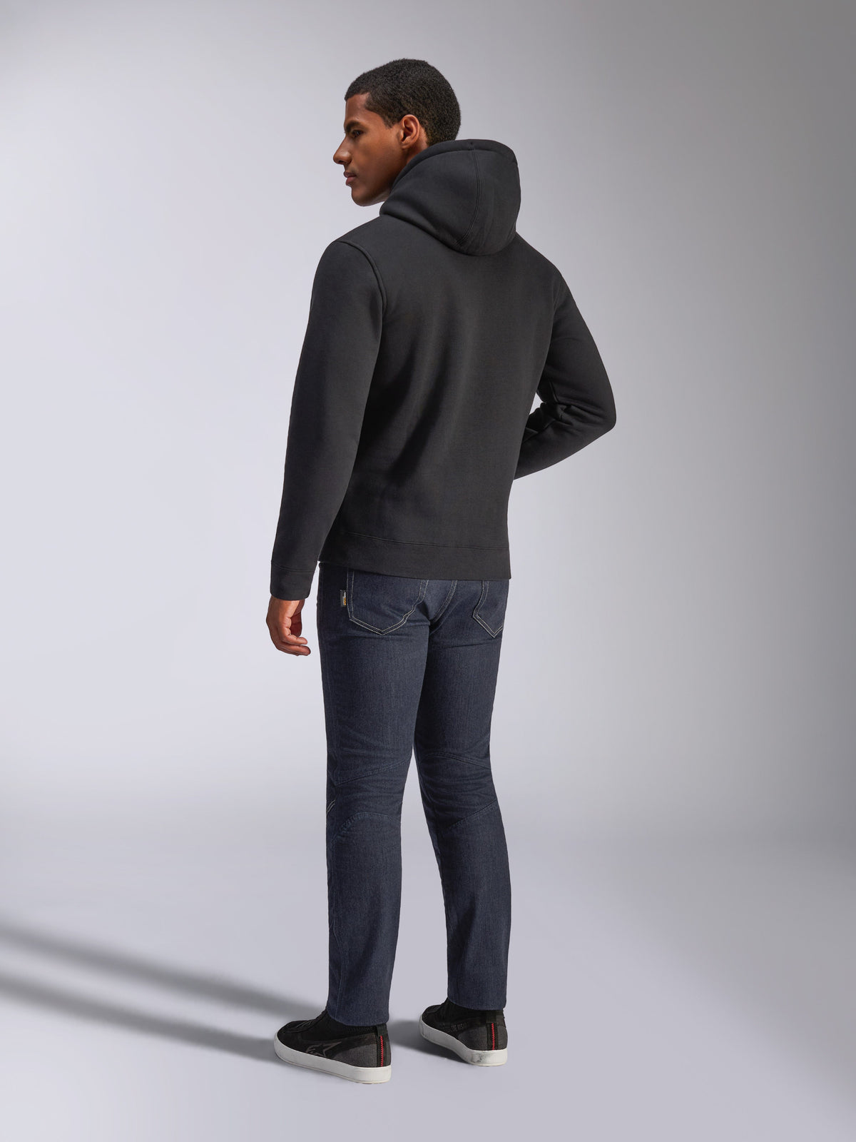 Alpinestars Blaze V3 Hoodie, widok z tyłu na mężczyznę w czarnej bluzie z kapturem, ciemne dżinsy i czarne sneakersy, casualowa odzież uliczna inspirowana motorsportem