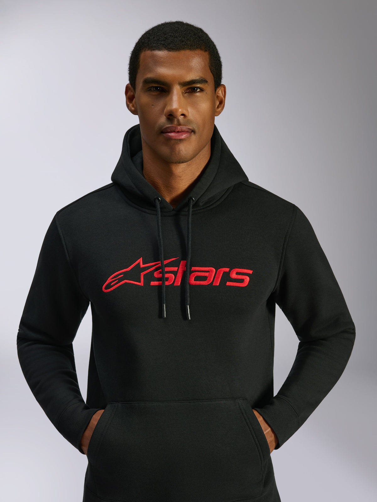 Alpinestars Blaze V3 Hoodie, czarna bluza z kapturem, czerwone logo Alpinestars na klatce piersiowej, noszona przez mężczyznę, kaptur ze sznurkiem, długie rękawy, nowoczesny design inspirowany motorsportem