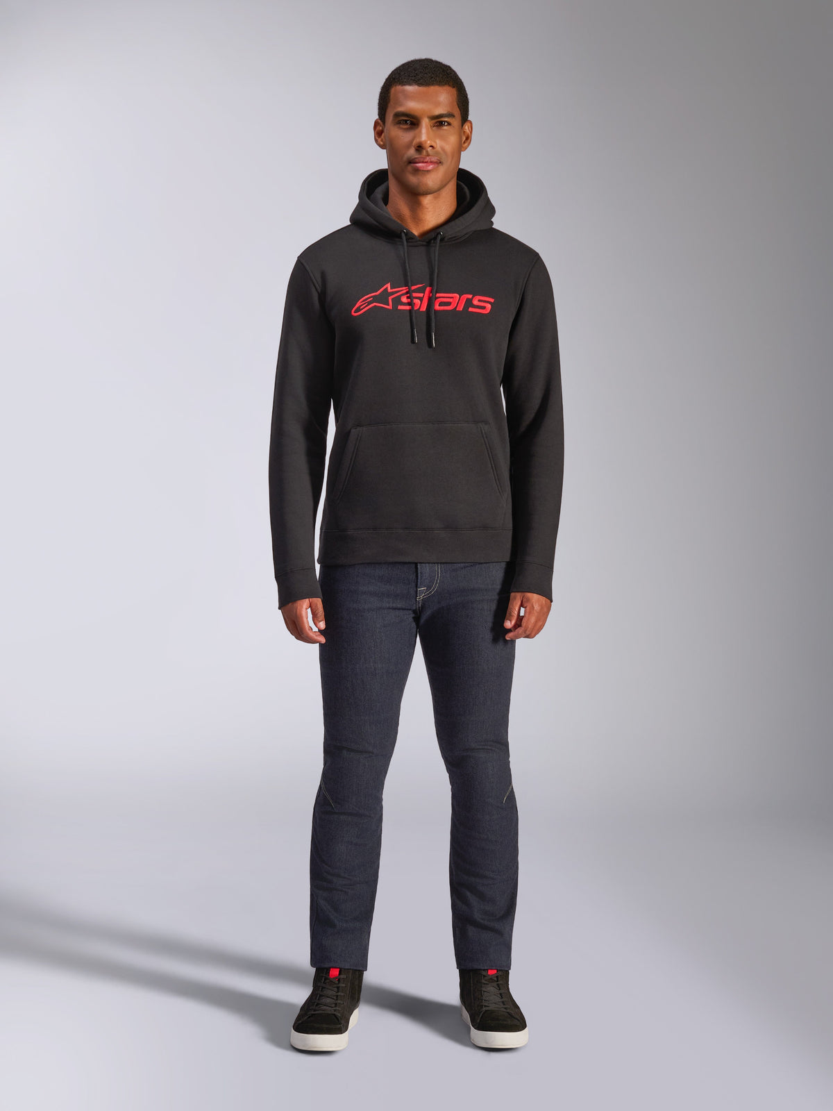 Bluza z kapturem Alpinestars Blaze V3, casual streetwear, czarna z czerwonym logo Astars, widok z przodu, prezentowana przez modela w ciemnych jeansach i czarnych sneakersach, nowoczesny polarowy design inspirowany motorsportem