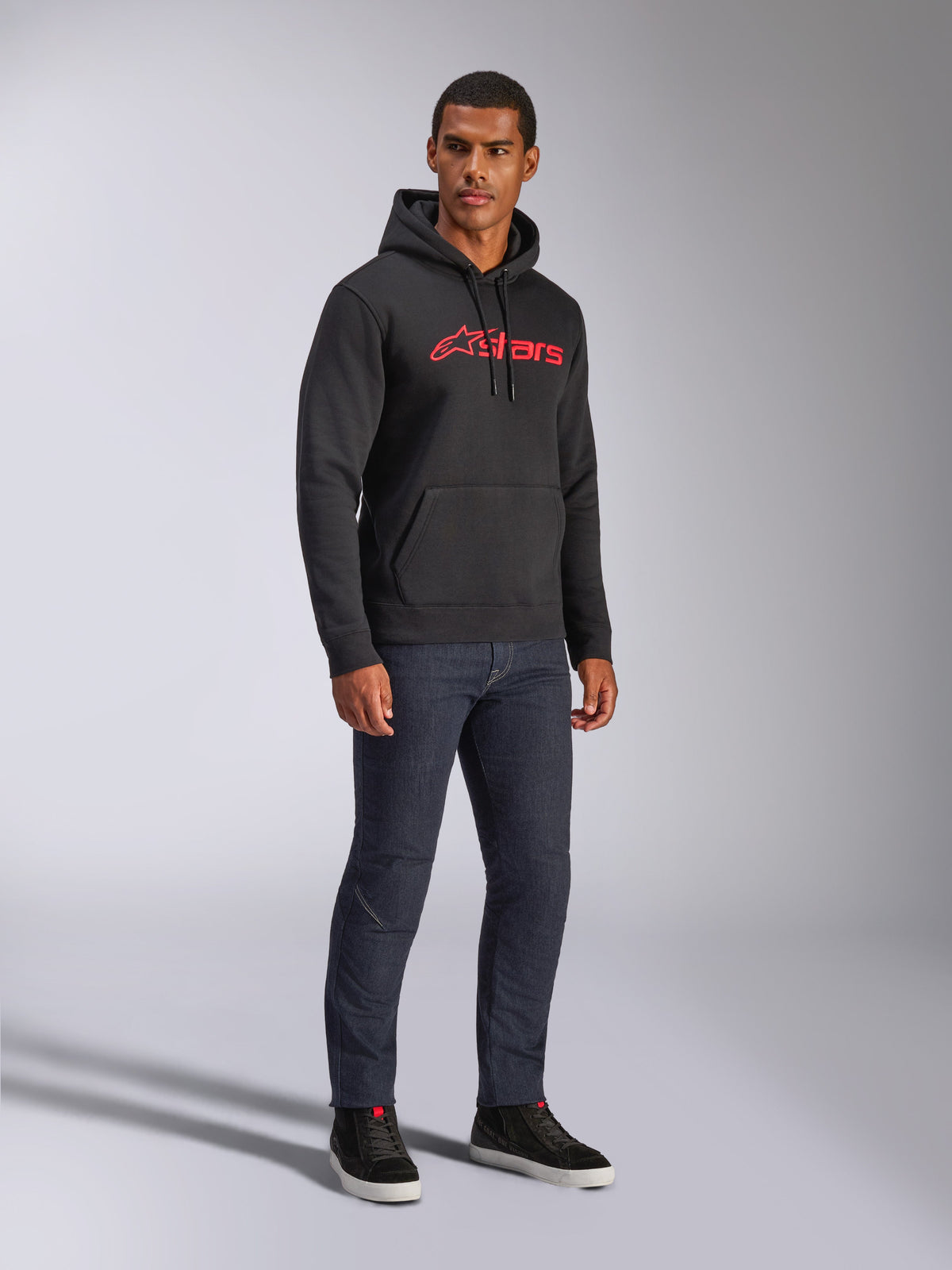 Alpinestars Blaze V3 Hoodie, czarna bluza polarowa z kapturem z czerwonym logo Alpinestars na piersi, noszona przez modela w ciemnych jeansach i czarnych sneakersach, codzienna odzież uliczna inspirowana motorsportem