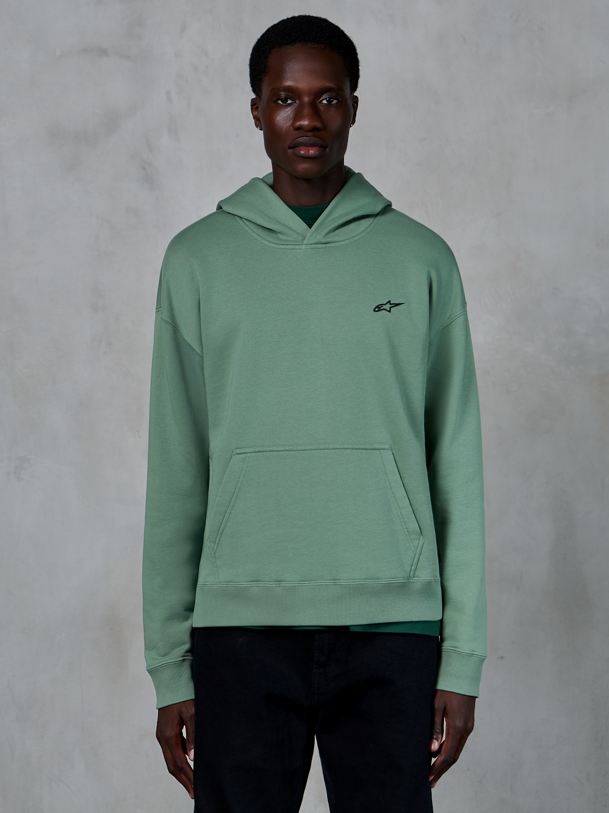 Alpinestars Essentials Ageless Hoodie, zielona bluza z kapturem (Cactus), lifestylowa bluza z polaru z wykończeniem stone-wash, małe czarne logo gwiazdy na piersi, przednia kieszeń typu kangurka, model prezentujący produkt w minimalistycznym otoczeniu