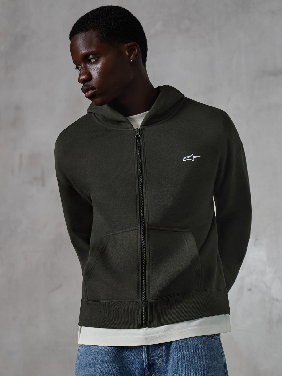 Alpinestars Essentials Ageless Zip Hoodie, casualowa bluza streetwear w kolorze Off Black, rozpinany polar z małym białym logo Alpinestars na piersi, noszona przez modela w niebieskich dżinsach, luźny krój z przednimi kieszeniami i wykończeniem stone-washed