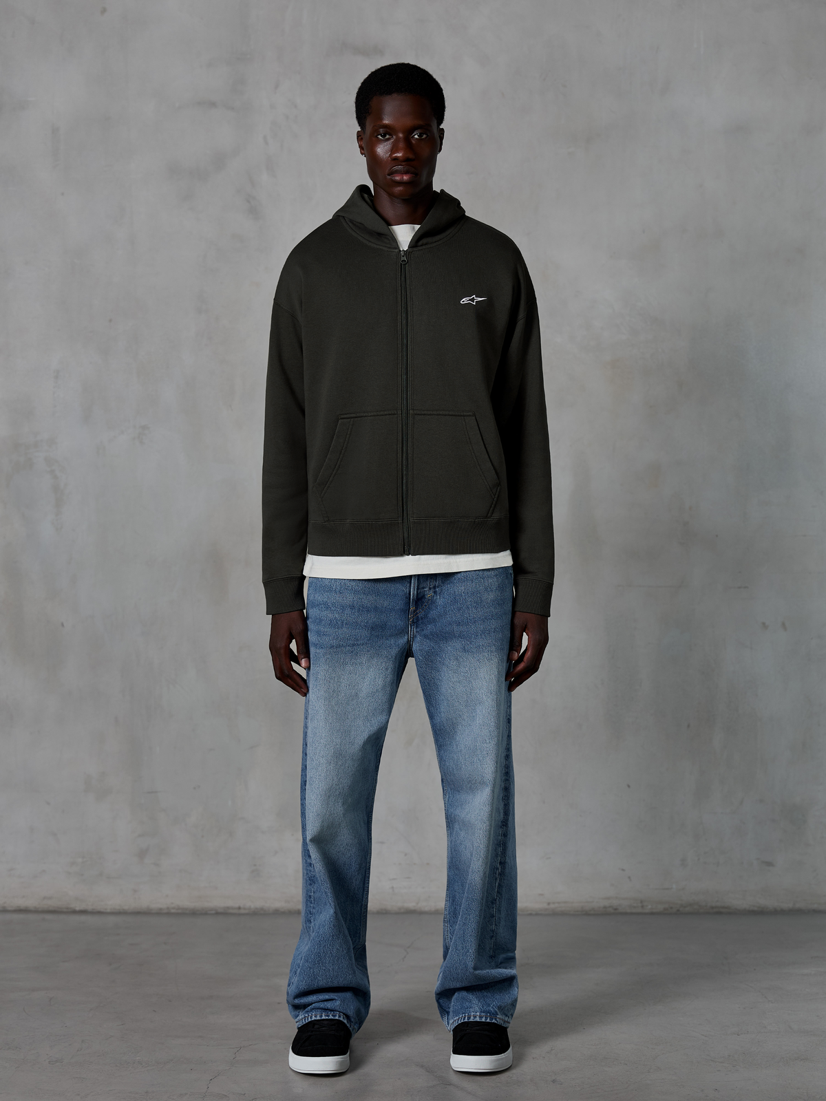 Alpinestars Essentials Ageless Zip Hoodie, rozpinana bluza z kapturem, kolor Off Black, z małym białym logo Alpinestars na piersi, na modelu w białym t-shircie i niebieskich jeansach, luźny krój, casualowa odzież motorsportowa