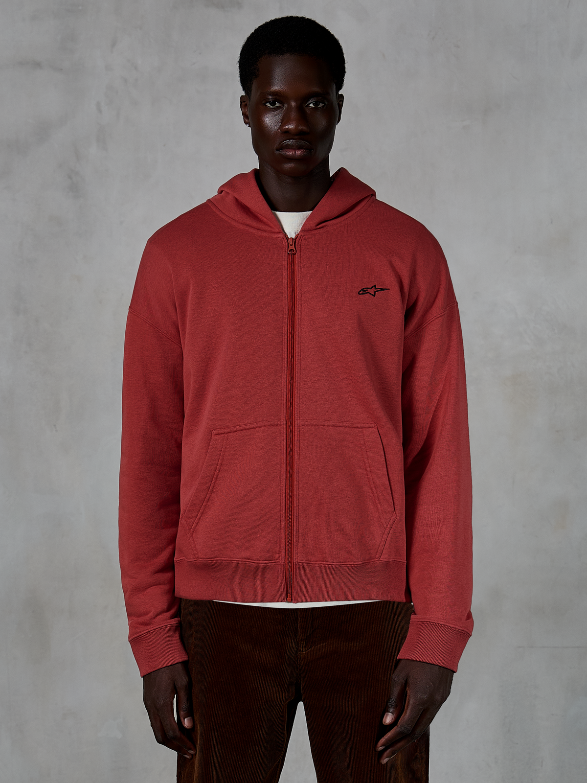 Alpinestars Essentials Ageless Zip Hoodie, ceglastoczerwona bluza z kapturem zapinana na zamek, noszona przez modela z brązowymi sztruksowymi spodniami, małe czarne logo na piersi, luźny fason w stylu streetwear