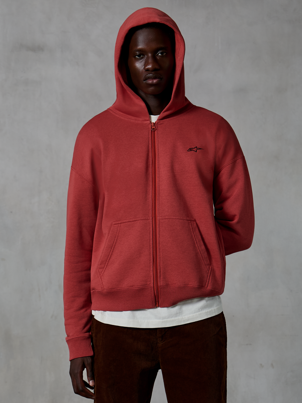 Alpinestars Essentials Ageless Zip Hoodie, sportowa bluza w kolorze ceglastym, fason z pełnym zamkiem i założonym kapturem, małe czarne logo na piersi, model w białym t-shircie i brązowych sztruksowych spodniach, szare tło