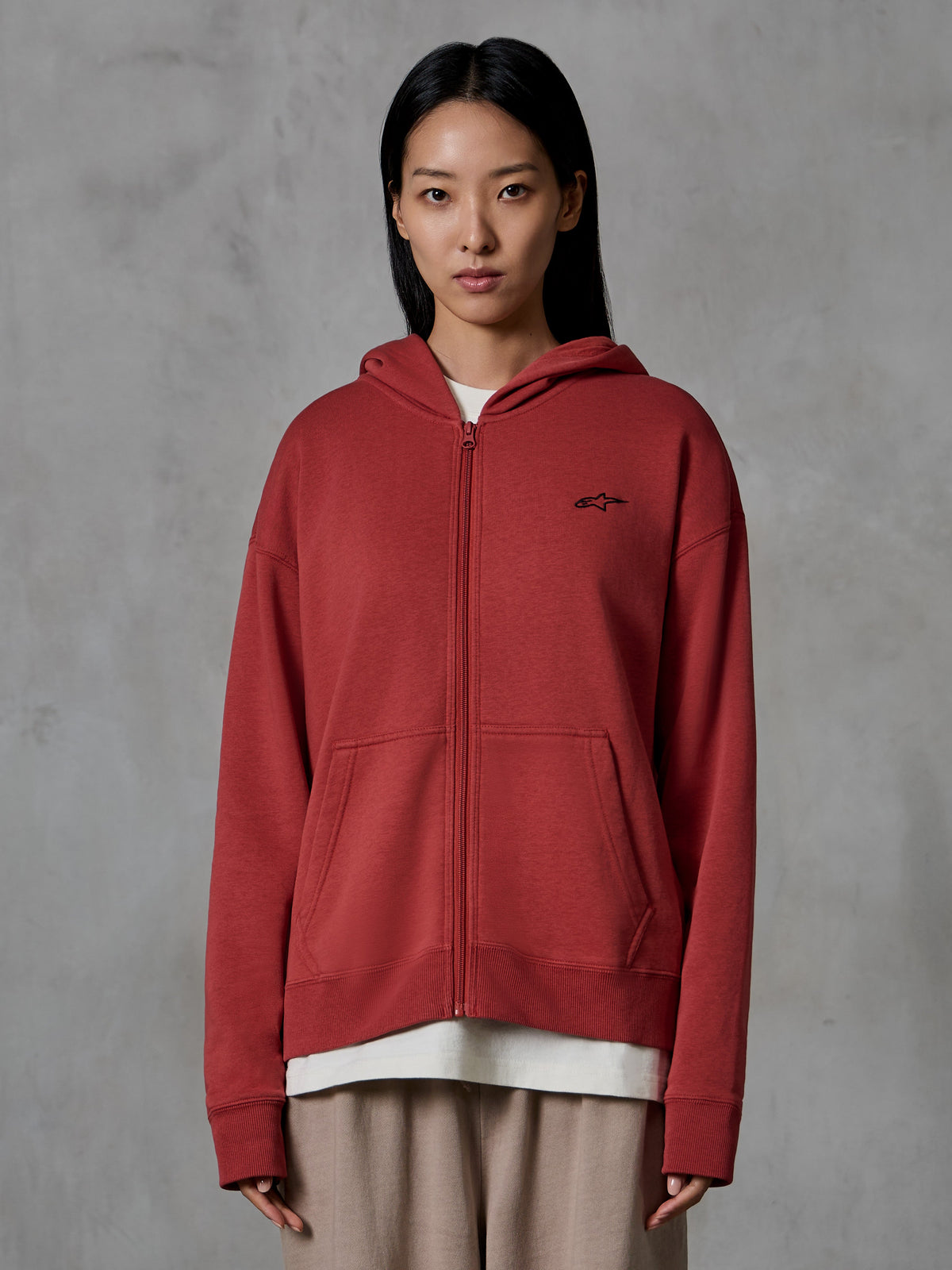 Alpinestars Essentials Ageless Zip Hoodie, kolor ceglastoczerwony, odzież streetwearowa, prezentowana na modelce, model rozpinany z kapturem, małe czarne logo Alpinestars na piersi, luźny krój, kompaktowy polar