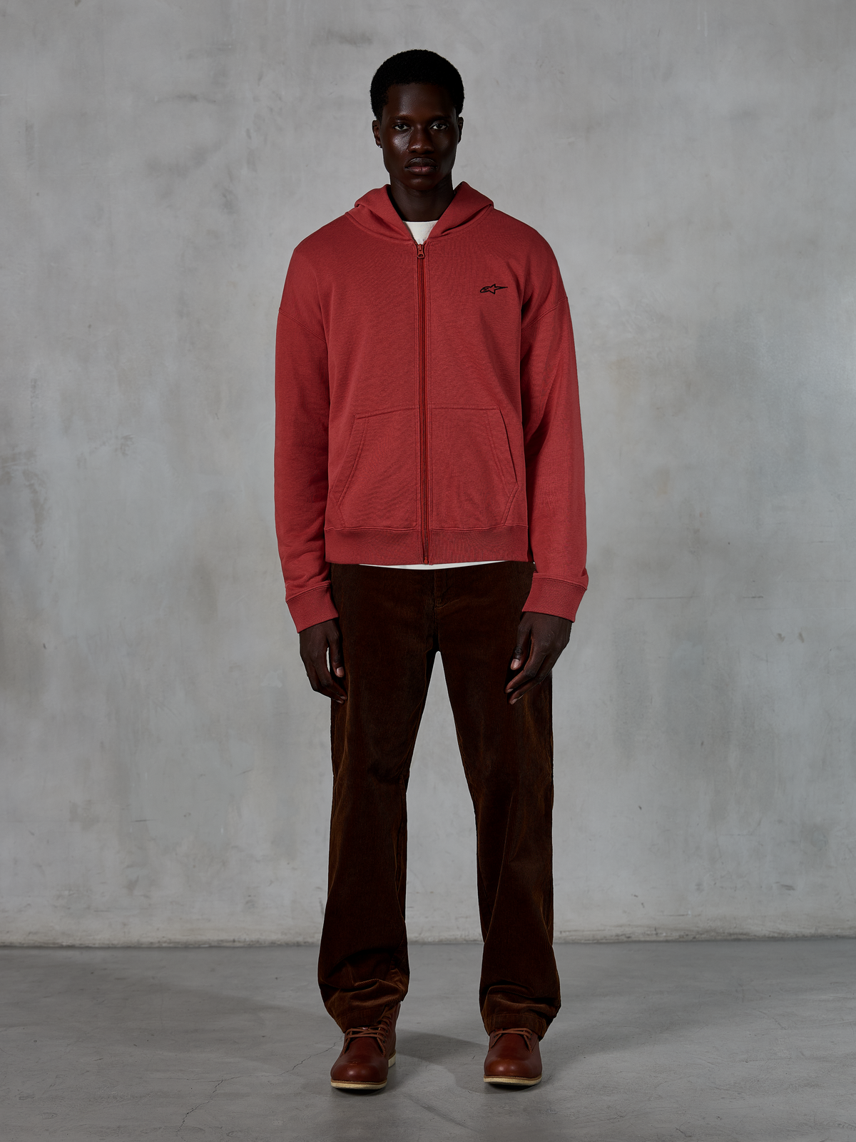 Bluza z kapturem Alpinestars Essentials Ageless Zip, casualowy streetwear, ceglasta czerwień z małym czarnym logo gwiazdy, zamek na całej długości, prezentowana przez modela w brązowych sztruksowych spodniach i butach, design premium inspirowany sportami motorowymi
