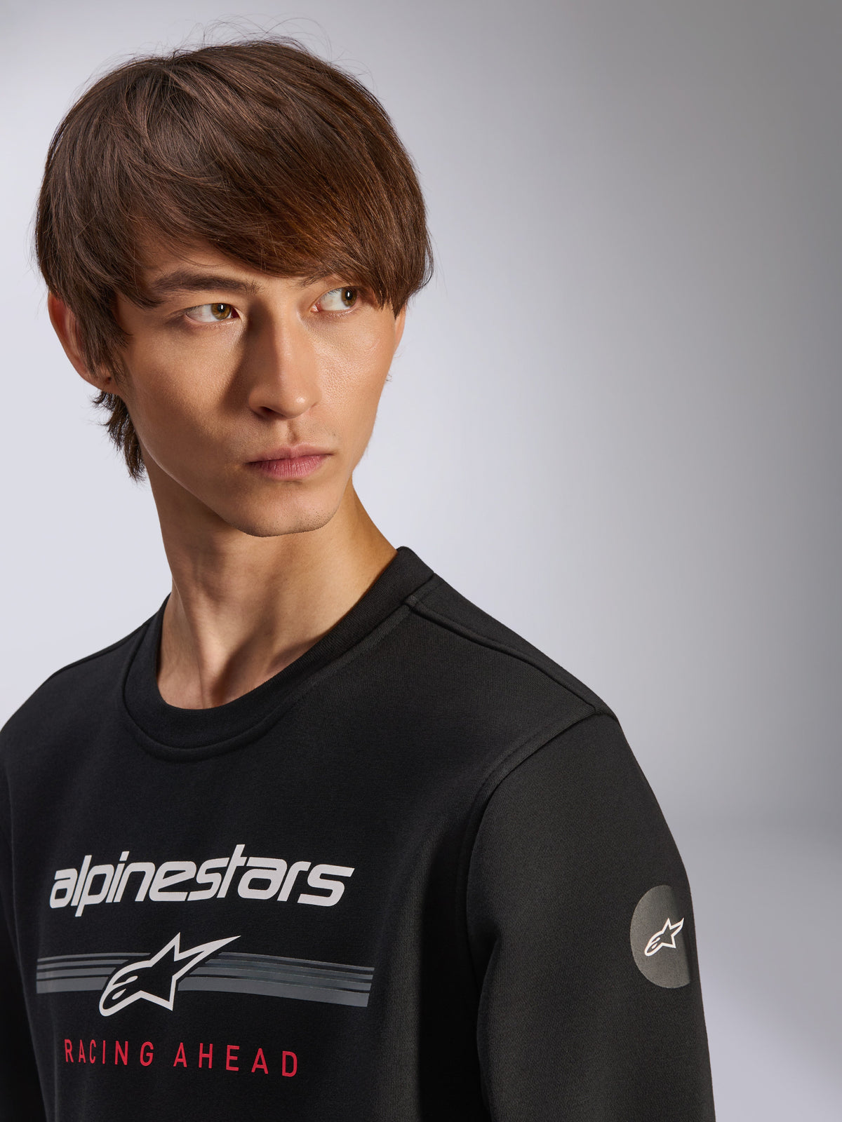 Alpinestars Bettern Crew Sweatshirt, black, featuring white Alpinestars logo with grey stripes and red 'RACING AHEAD' text, circular star logo on the sleeve, casual motorsport-inspired streetwear