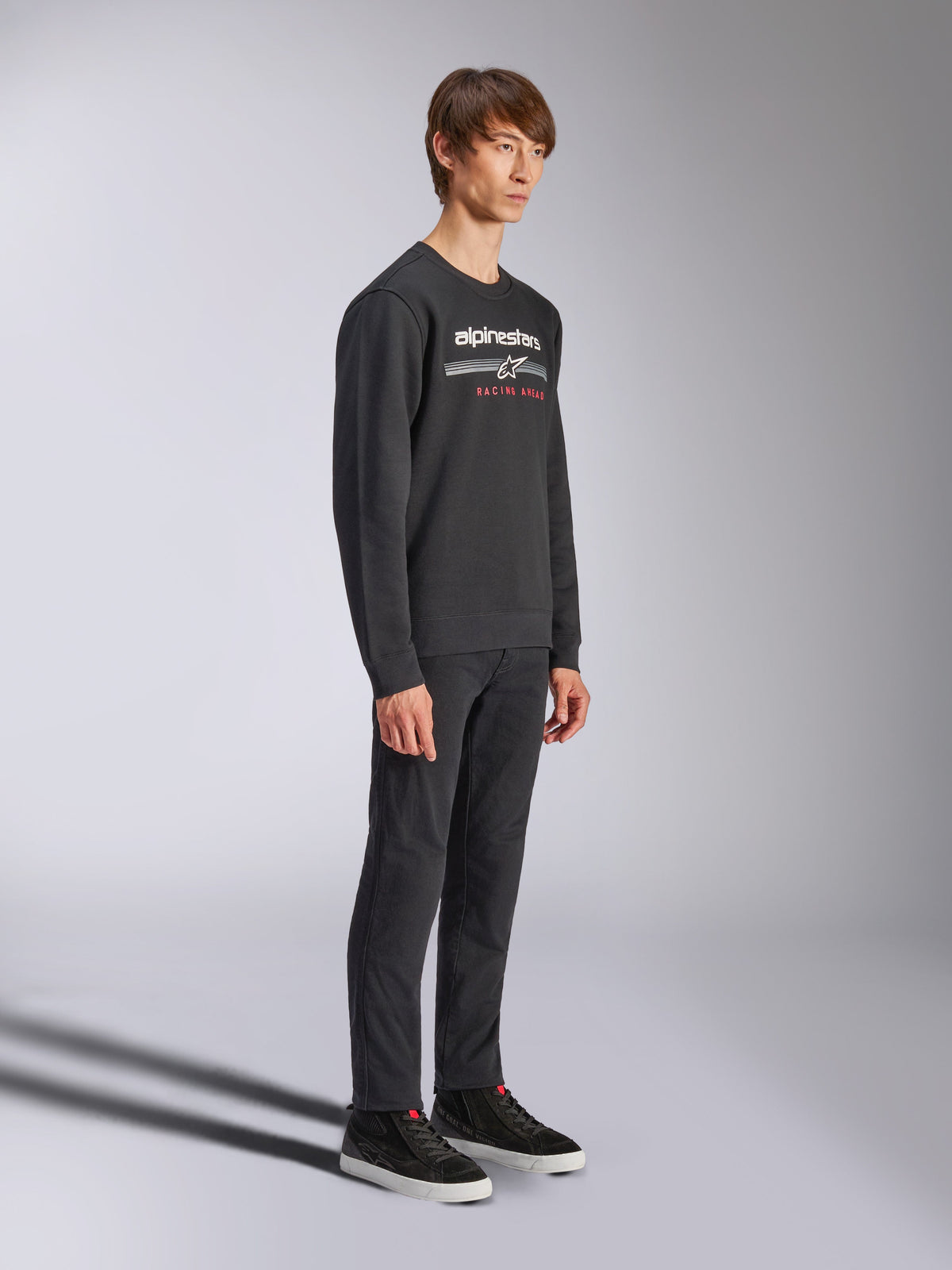 Alpinestars Bettern Crew Sweatshirt, black crewneck pullover, white alpinestars logo with 'RACING AHEAD' in red on the chest, paired with black pants and sneakers, casual motorsport-inspired streetwear