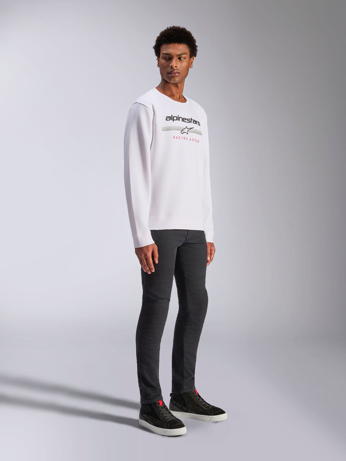 Alpinestars Bettern Crew, biała bluza typu crewneck z czarnym logo Alpinestars i czerwonym napisem „Racing Ahead”, casualowa odzież inspirowana motorsportem, prezentowana przez modela w ciemnych spodniach i czarnych sneakersach
