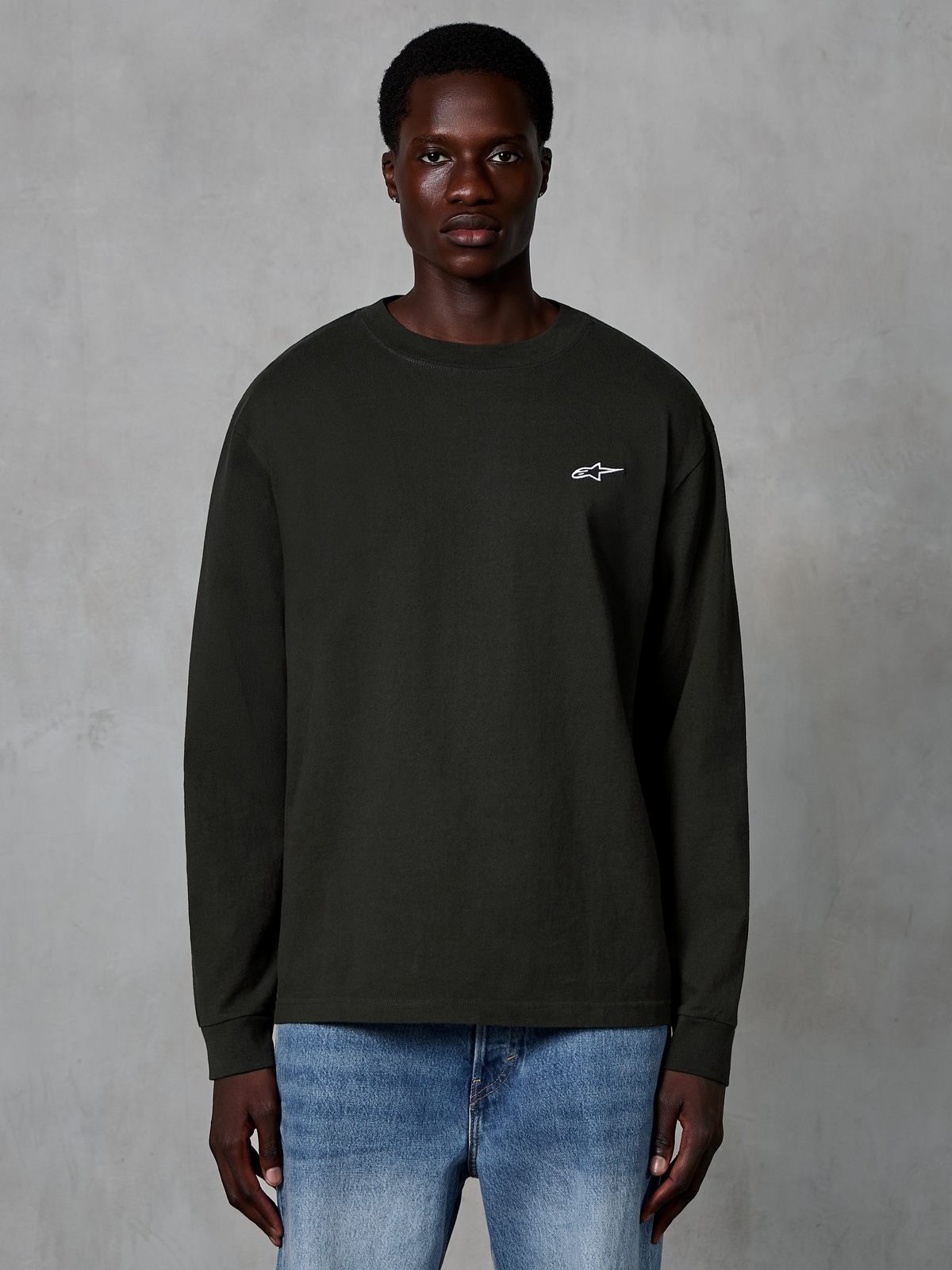 Alpinestars Essentials Ageless LS Tee, koszulka z długim rękawem, kolor Off Black, małe białe logo Alpinestars na piersi, noszona przez modela z niebieskimi jeansami, codzienna odzież streetwear z bawełny organicznej