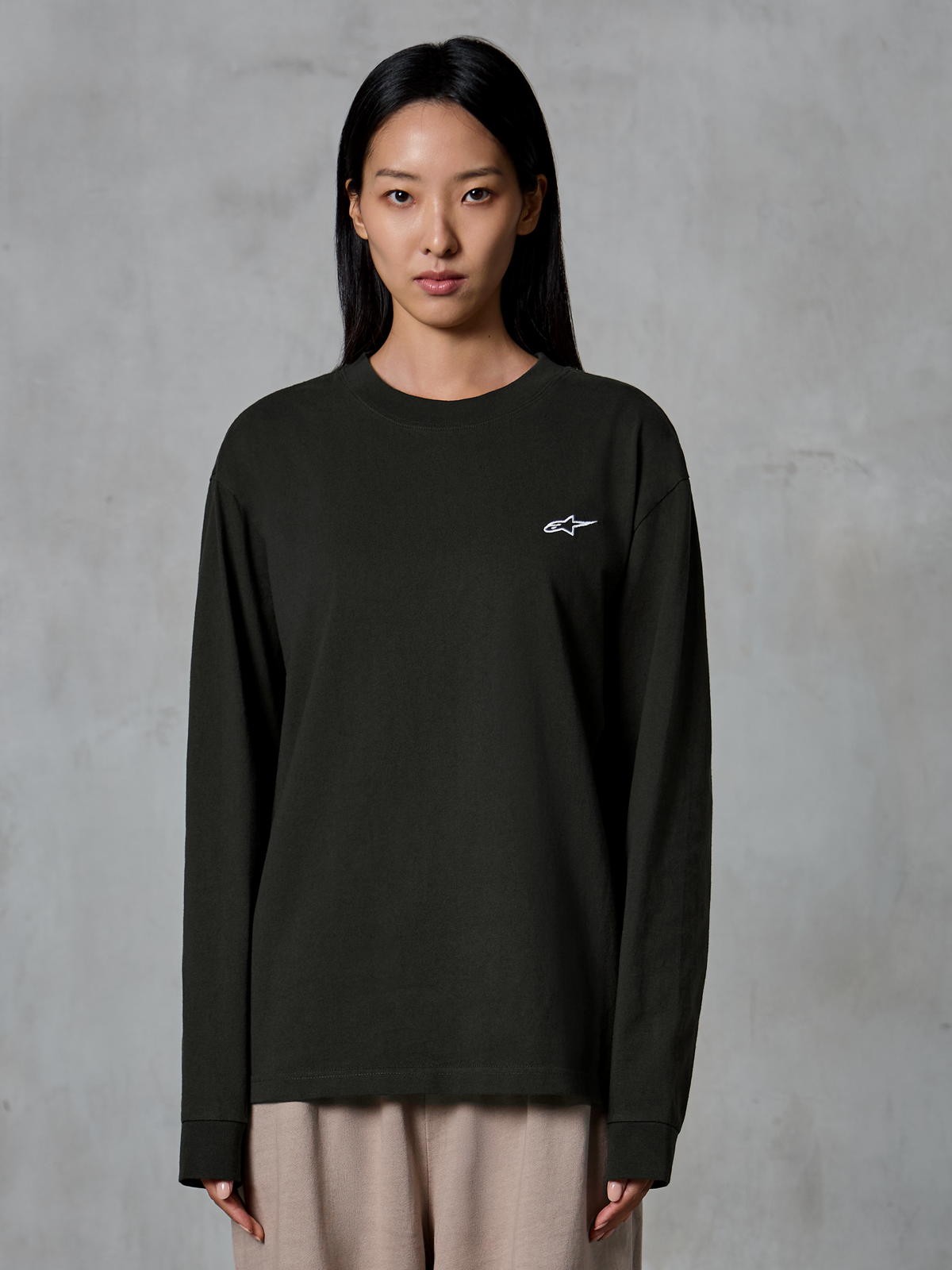 Modelka w koszulce Alpinestars Essentials Ageless LS Tee, t-shirt z długim rękawem, kolor Off Black, małe białe logo Alpinestars na piersi, beżowe spodnie, casualowy styl streetwear
