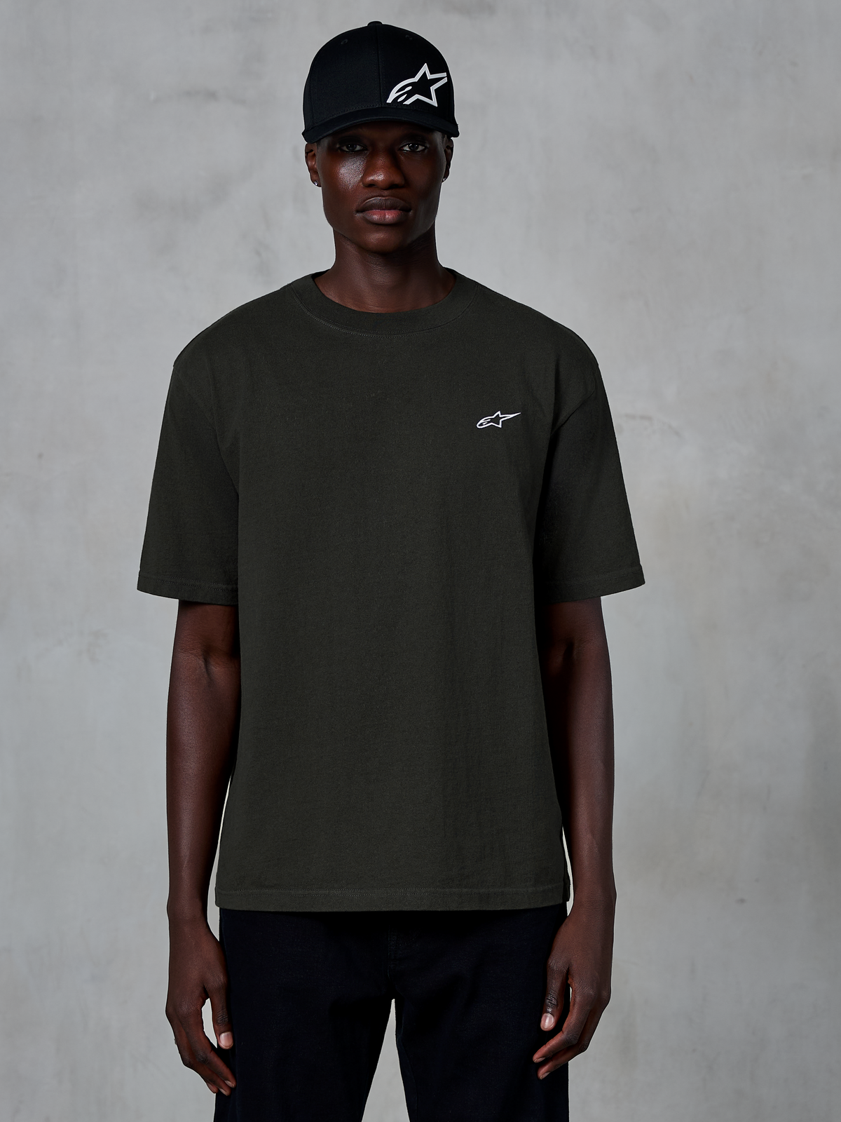 Alpinestars Essentials Ageless SS Tee, t-shirt z krótkim rękawem, kolor Off Black, noszony przez modela w czarnej czapce z logo, małe białe logo Alpinestars na piersi, codzienny streetwear inspirowany sportami motorowymi