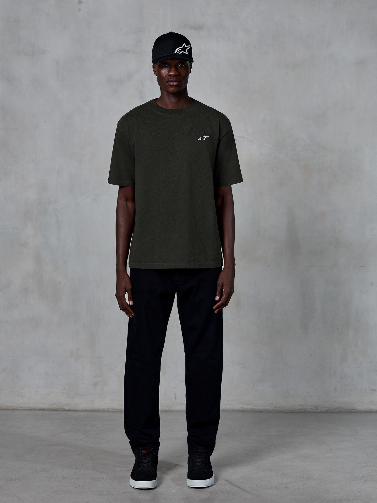 Alpinestars Essentials Ageless SS Tee, koszulka z krótkim rękawem, kolor Off Black, z małym białym logo Alpinestars na piersi, noszona przez modela w czarnej czapce i spodniach, minimalistyczny styl lifestylowy