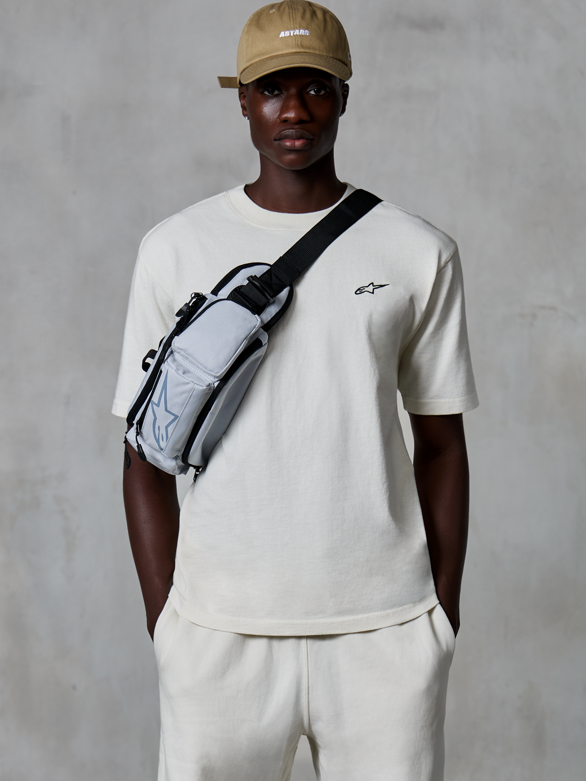 Alpinestars Essentials Ageless SS Tee, Short Sleeve T-Shirt, Off White, worn with matching shorts and a tan cap, featuring a small black logo on the chest and a grey crossbody bag, casual motorsport-inspired streetwear